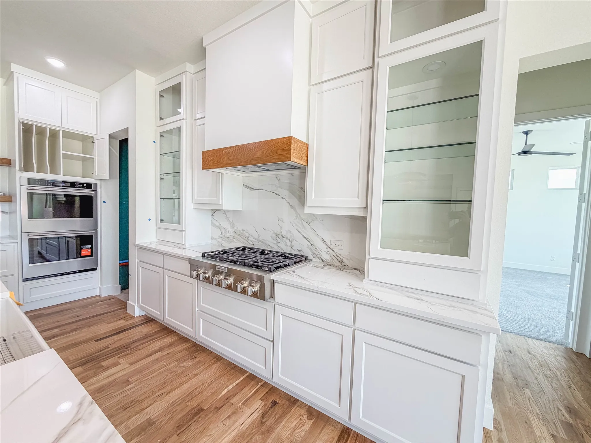 White cabinetry with glass-front display cabinets, light wood-finish flooring, stainless steel gas range with a matching range hood featuring a wood accent, and integrated stainless steel double oven