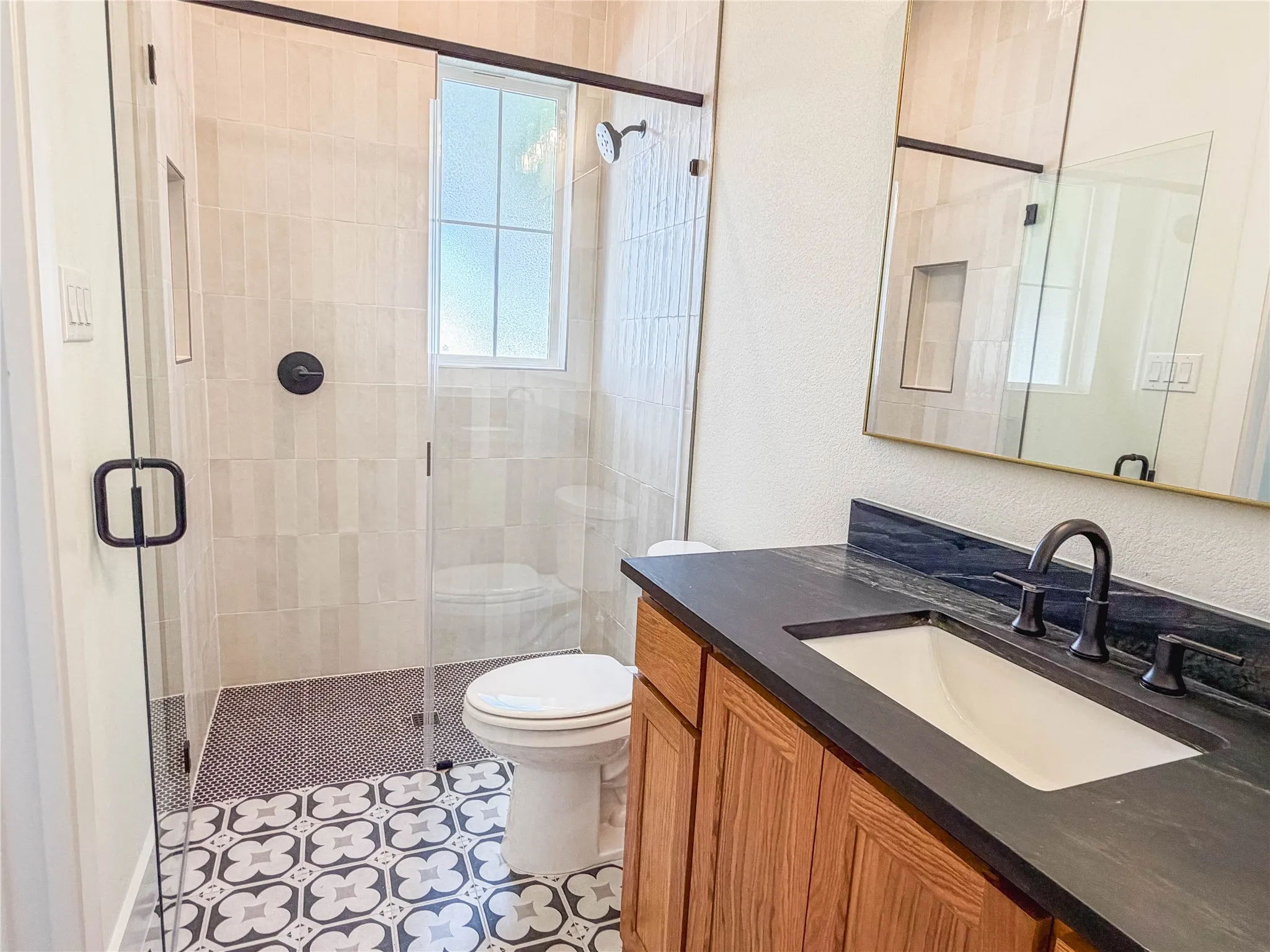 Walk-in shower with clear glass enclosure and black hardware, patterned floor tile, single vanity with dark countertop, and rectangular undermount sink