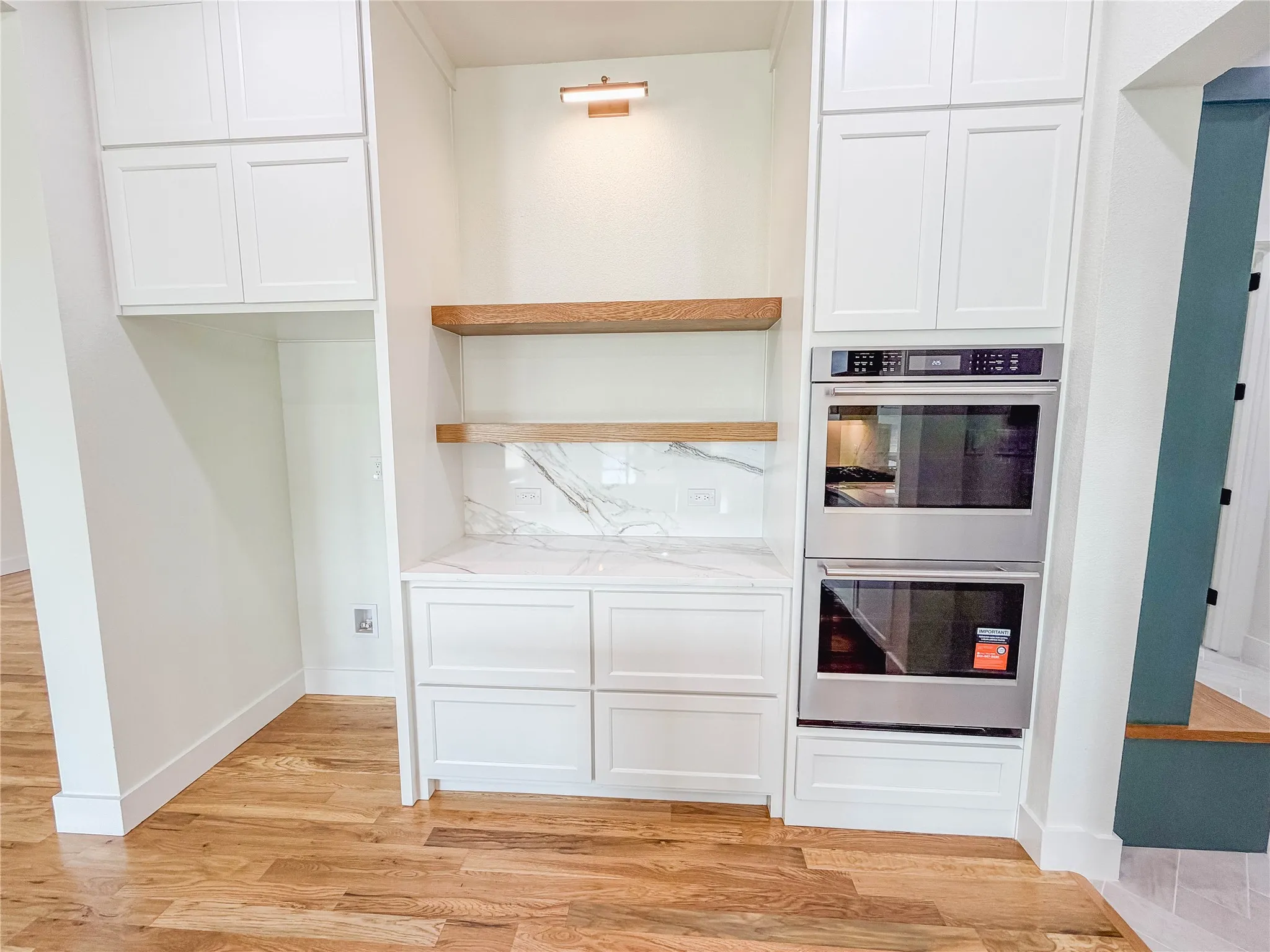 White shaker cabinetry, wood-finish flooring, built-in double ovens, marble-look backsplash, and floating wood shelves