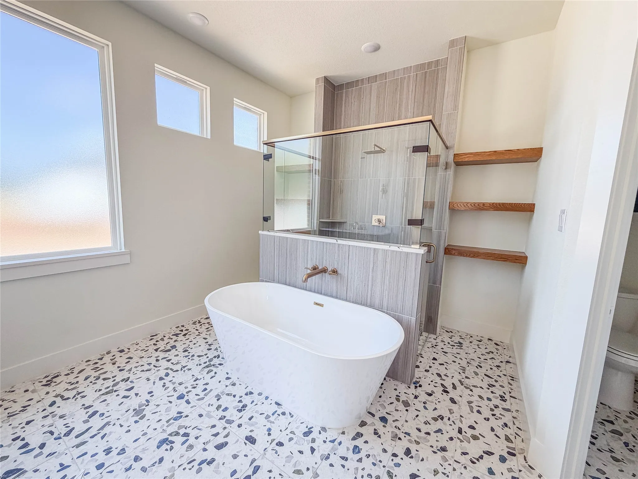 Freestanding soaking tub with a wall-mounted bronze faucet, glass-enclosed shower with tile surround, wood-finish shelving, and patterned tile flooring