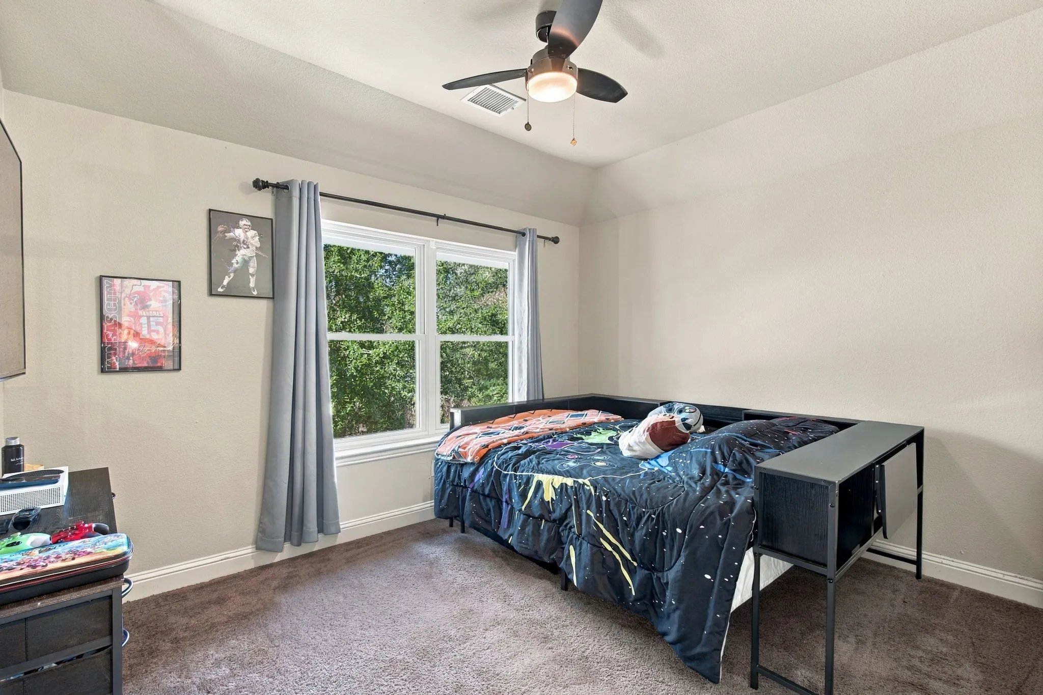 Comfortable upstairs bedroom featuring a large window with leafy views, ceiling fan, neutral walls, and carpeting.
