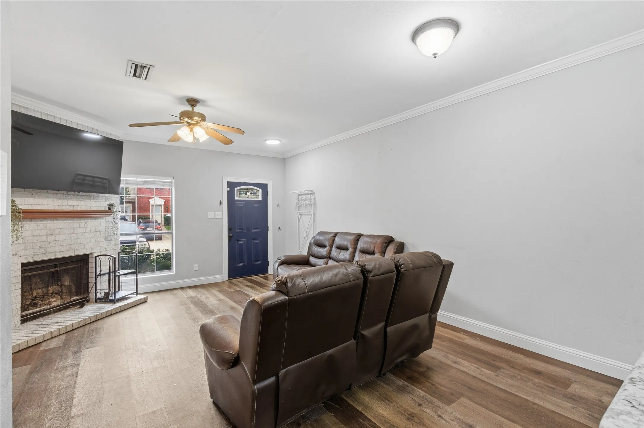 Living area with wood-finish flooring, a white brick fireplace, and a ceiling fan with integrated lighting