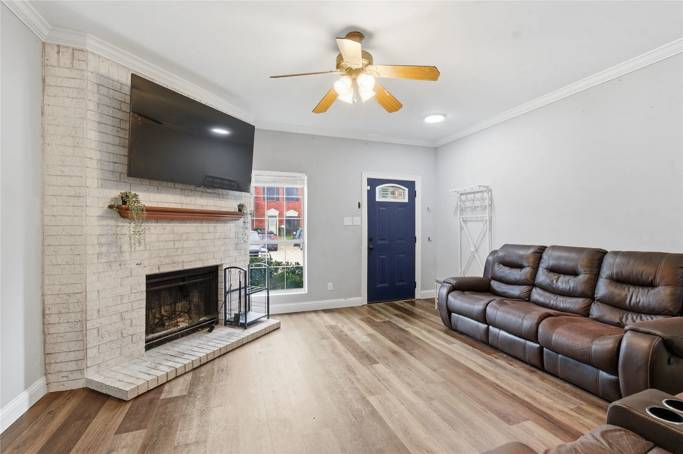 Corner fireplace with a wood mantel, wood-finish flooring, accent front door with decorative glass, and a ceiling fan with integrated lighting