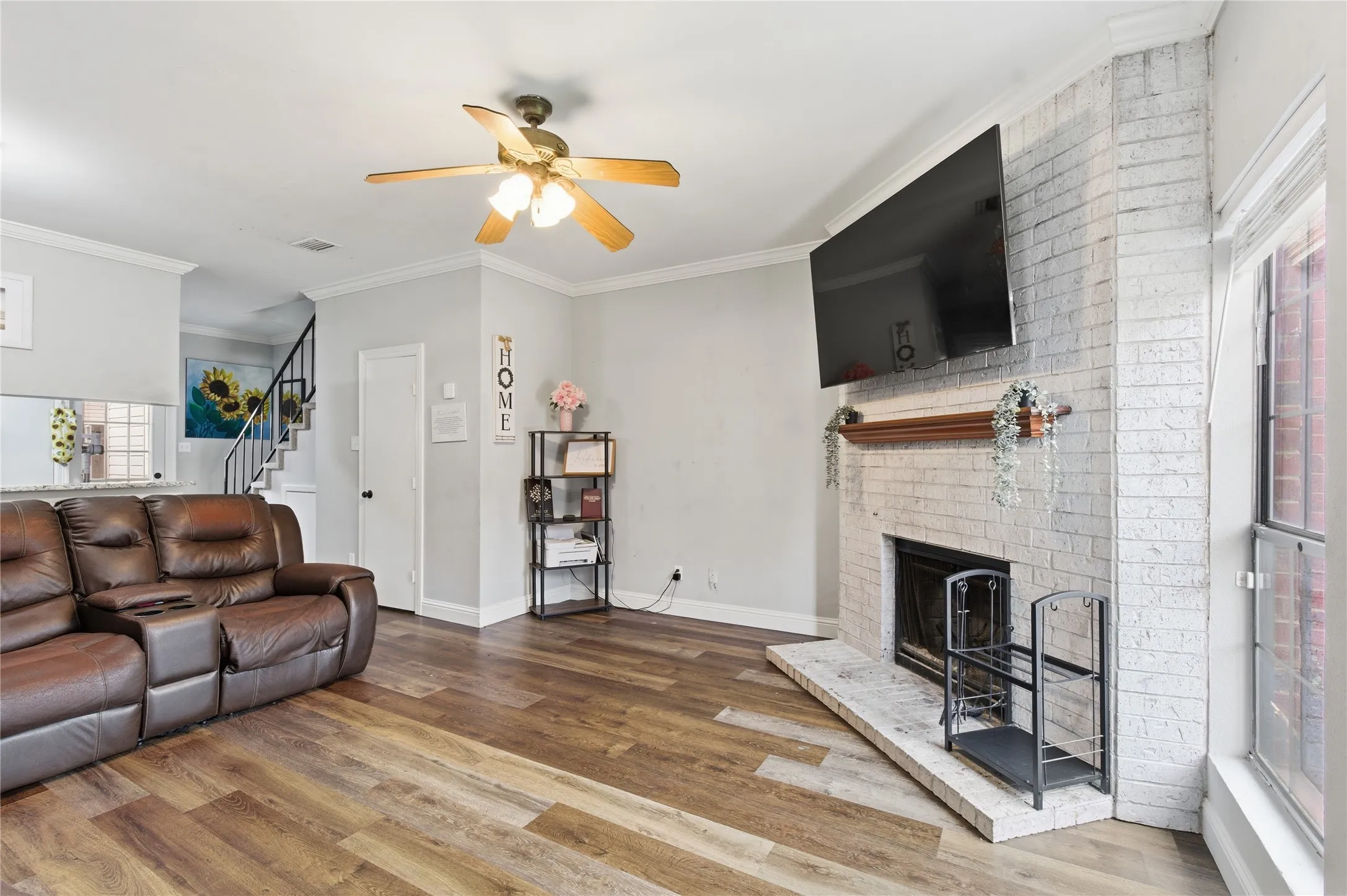 Living area featuring wood-finish flooring, a white-painted brick fireplace with a wood mantel, crown molding, and a ceiling fan with light fixture