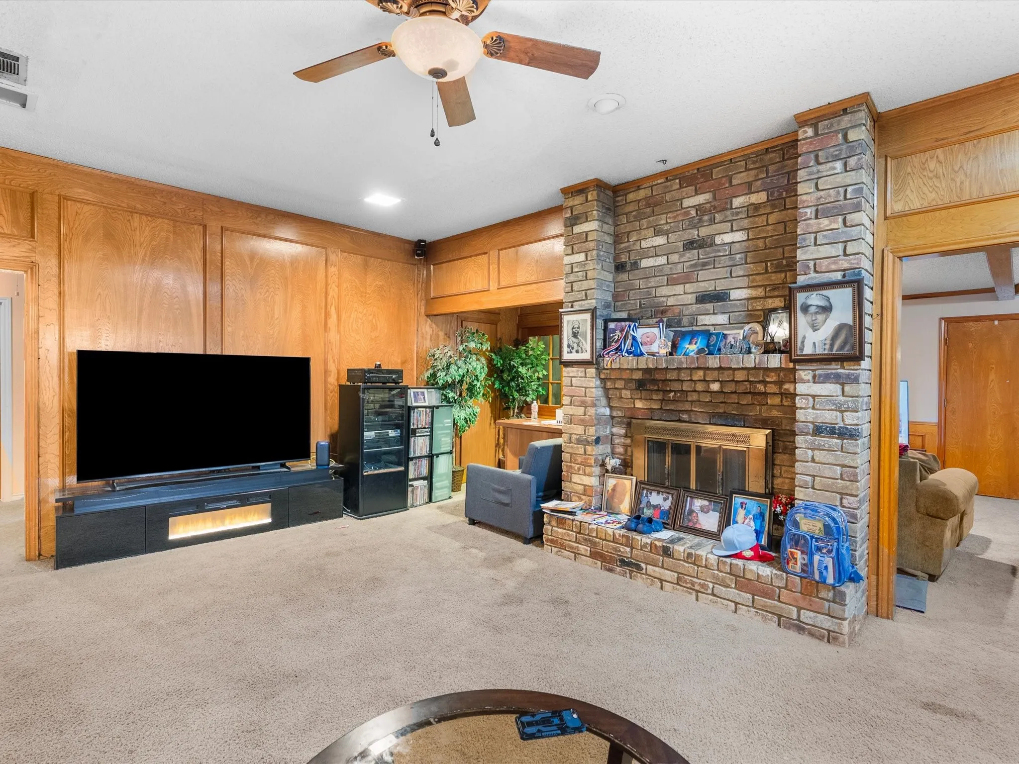 Brick fireplace with a brass-finish insert, wood paneling, and a ceiling fan