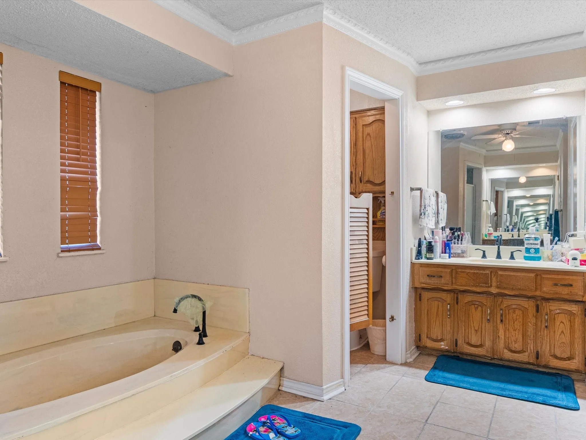 Built-in bathtub with tiered surround, wood-finish blinds, wood vanity with integrated sink, large wall mirror, and tile flooring
