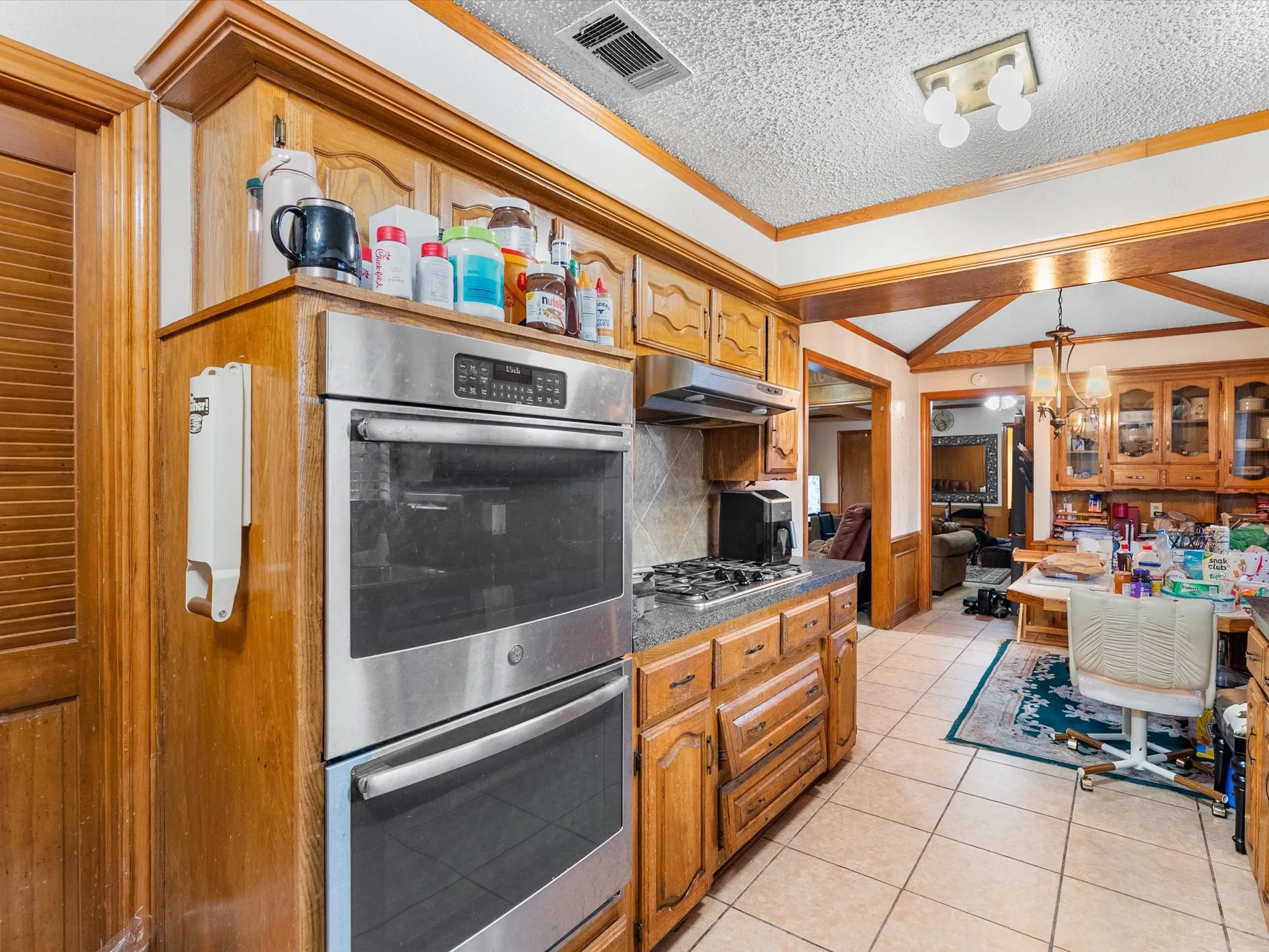 Double wall ovens, gas cooktop, wood cabinetry, tile flooring, and a ceiling-mounted light fixture