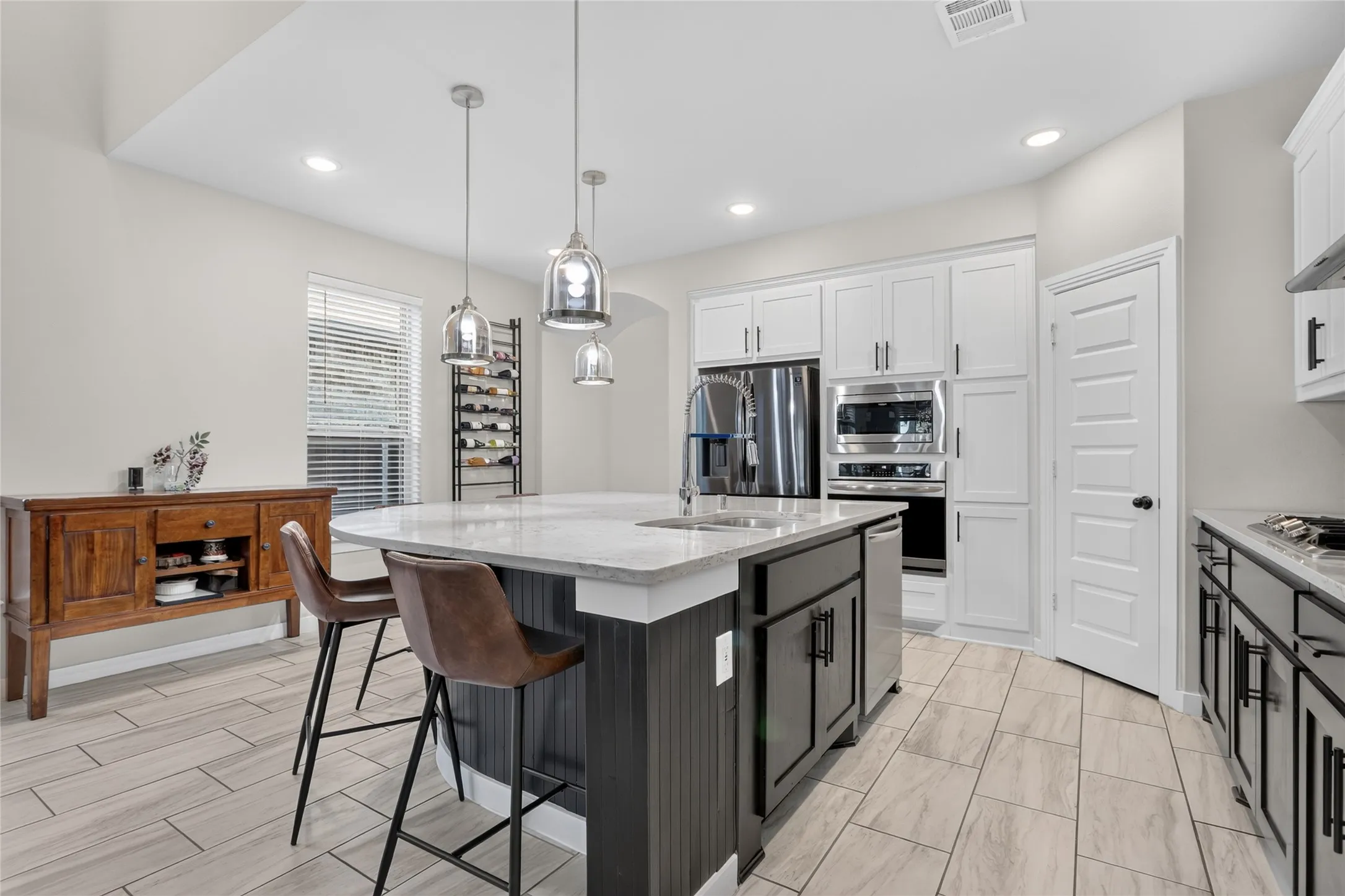 Kitchen featuring stainless steel appliances, white cabinetry, a central island with a sink, pendant lighting, and wood-finish tile flooring