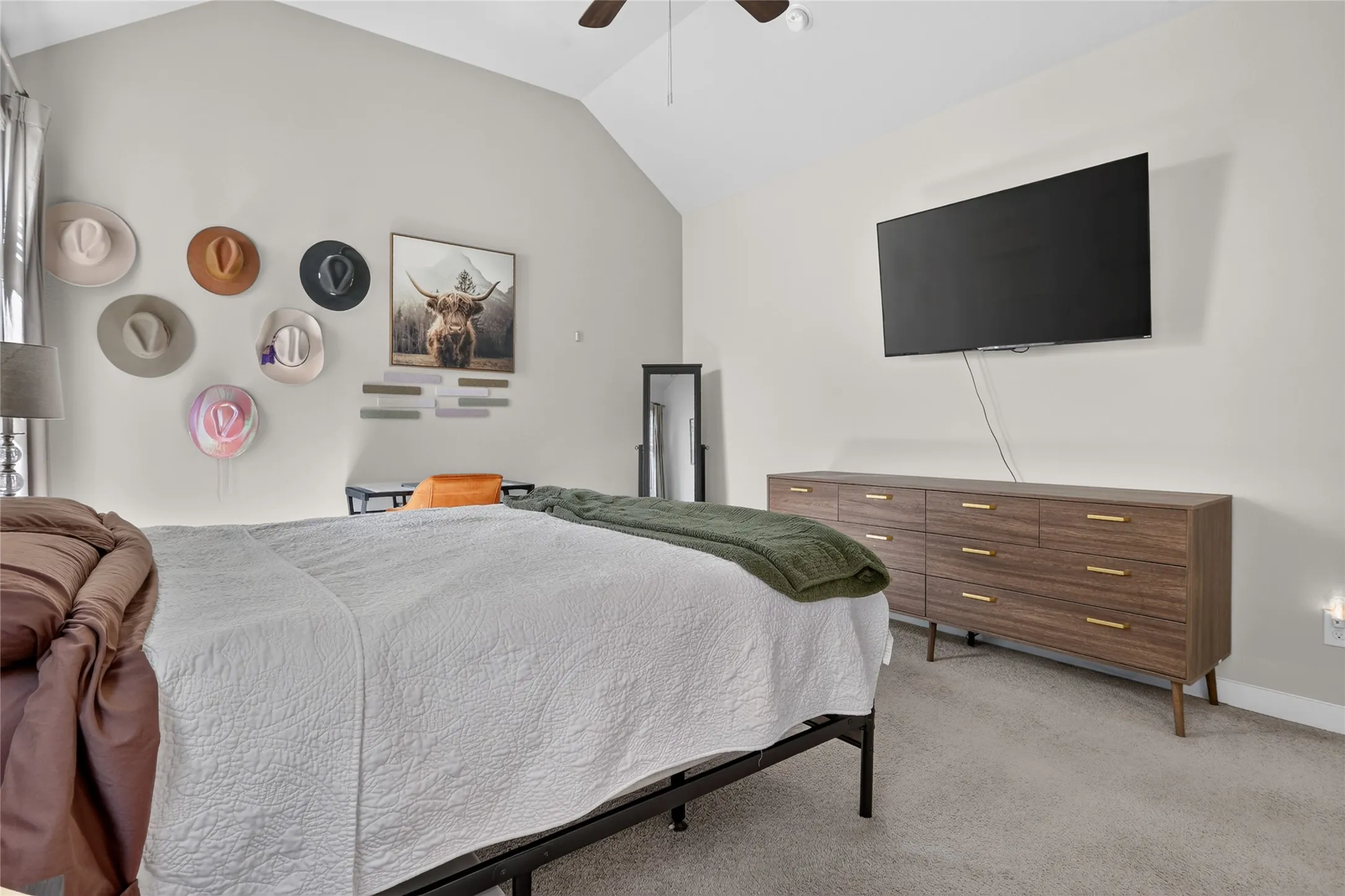 Vaulted ceiling, ceiling fan, wall-mounted television, wood-finish dresser with gold-tone hardware, and carpet flooring