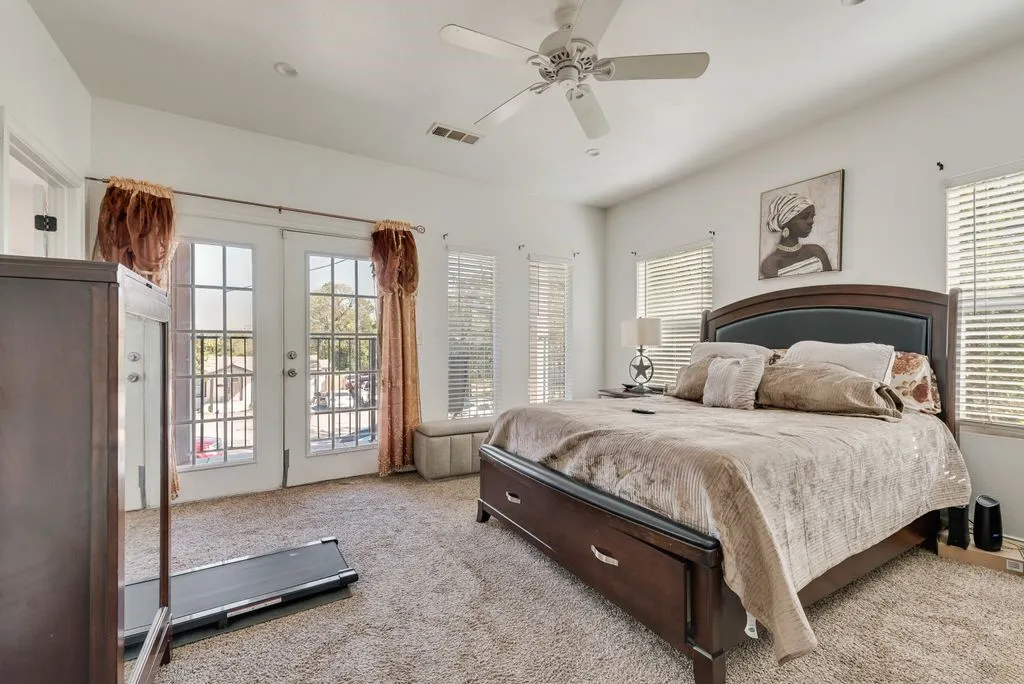 Spacious primary bedroom with French doors opening to a private balcony, ceiling fan, plantation shutters, wood-finish flooring, and carpet accents