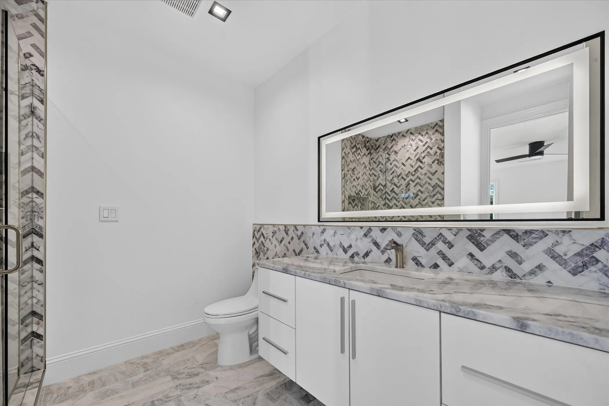 Modern bathroom featuring a glass-enclosed shower with chevron tile, an illuminated mirror, a vanity with a stone countertop, and wood-finish flooring