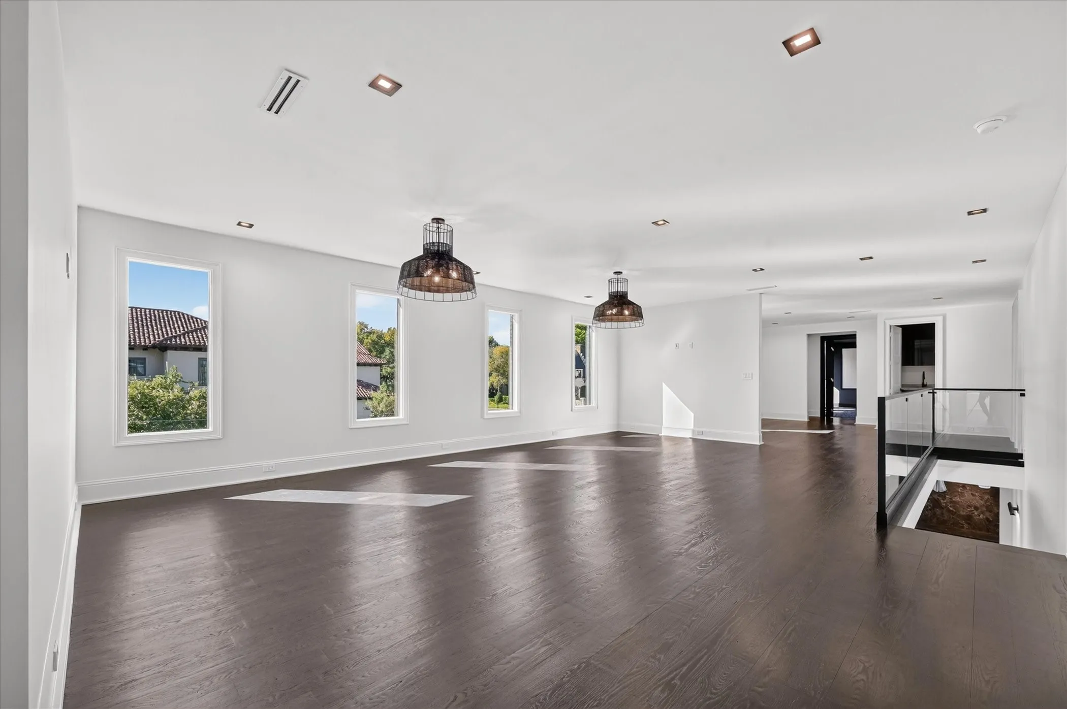 Dark wood-finish flooring, recessed ceiling lighting, three tall windows, two pendant light fixtures, and a glass-paneled railing
