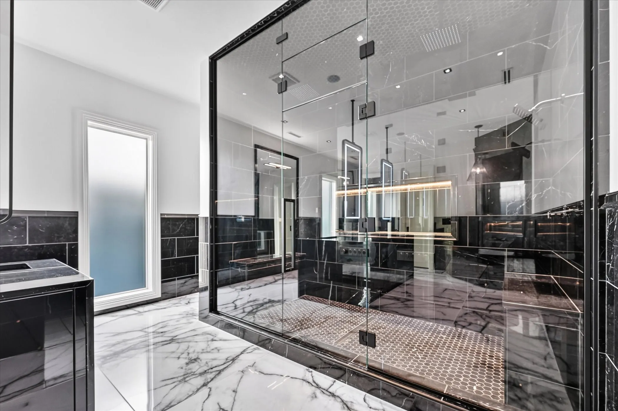 Spacious bathroom featuring white and black marble-style flooring, a large glass-enclosed shower with honeycomb tile, a frosted privacy window, and dark tiled wainscoting
