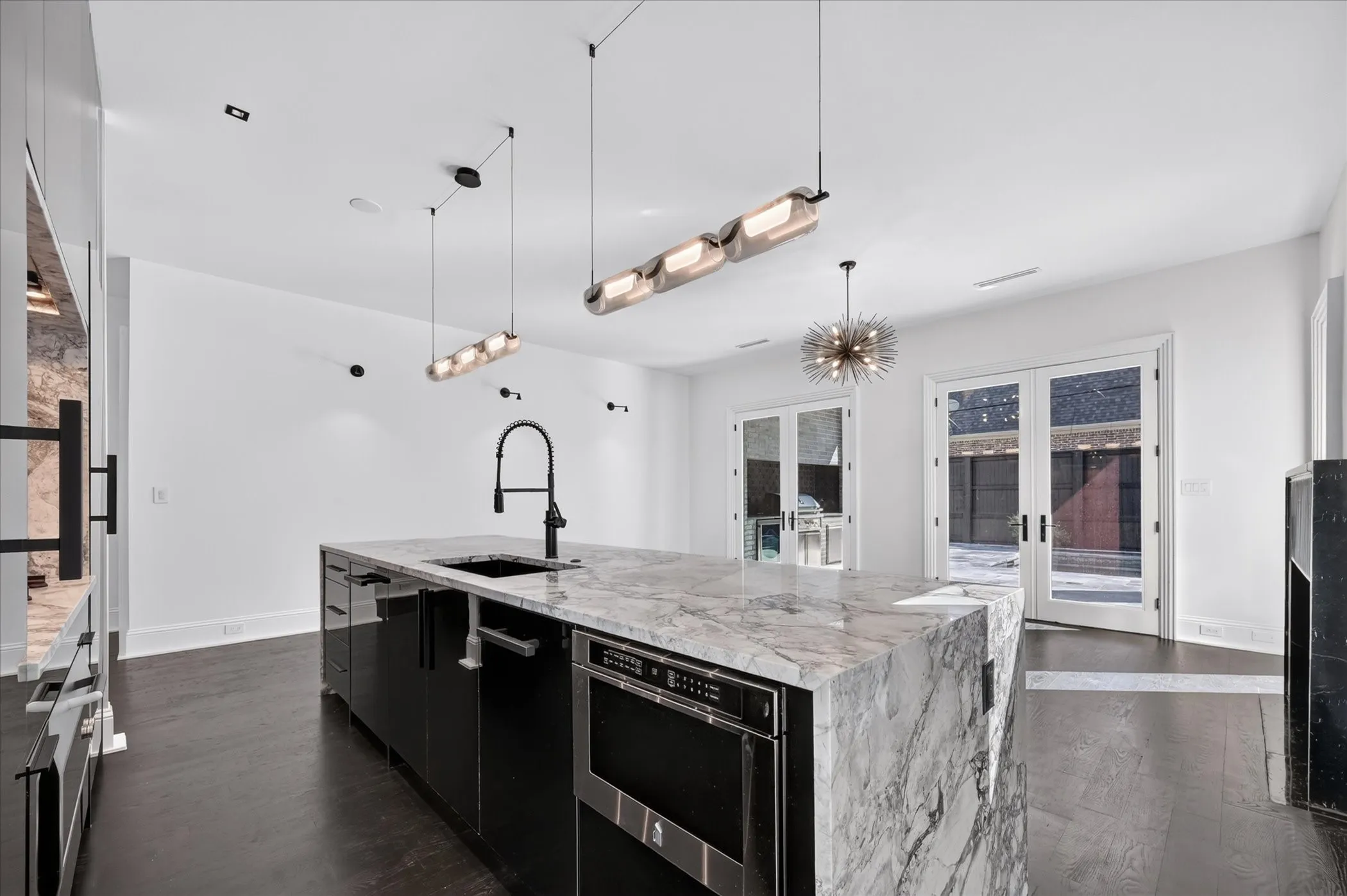 Gourmet kitchen featuring a waterfall island with integrated sink, sleek black cabinetry, dark wood-finish flooring, and multiple pendant light fixtures
