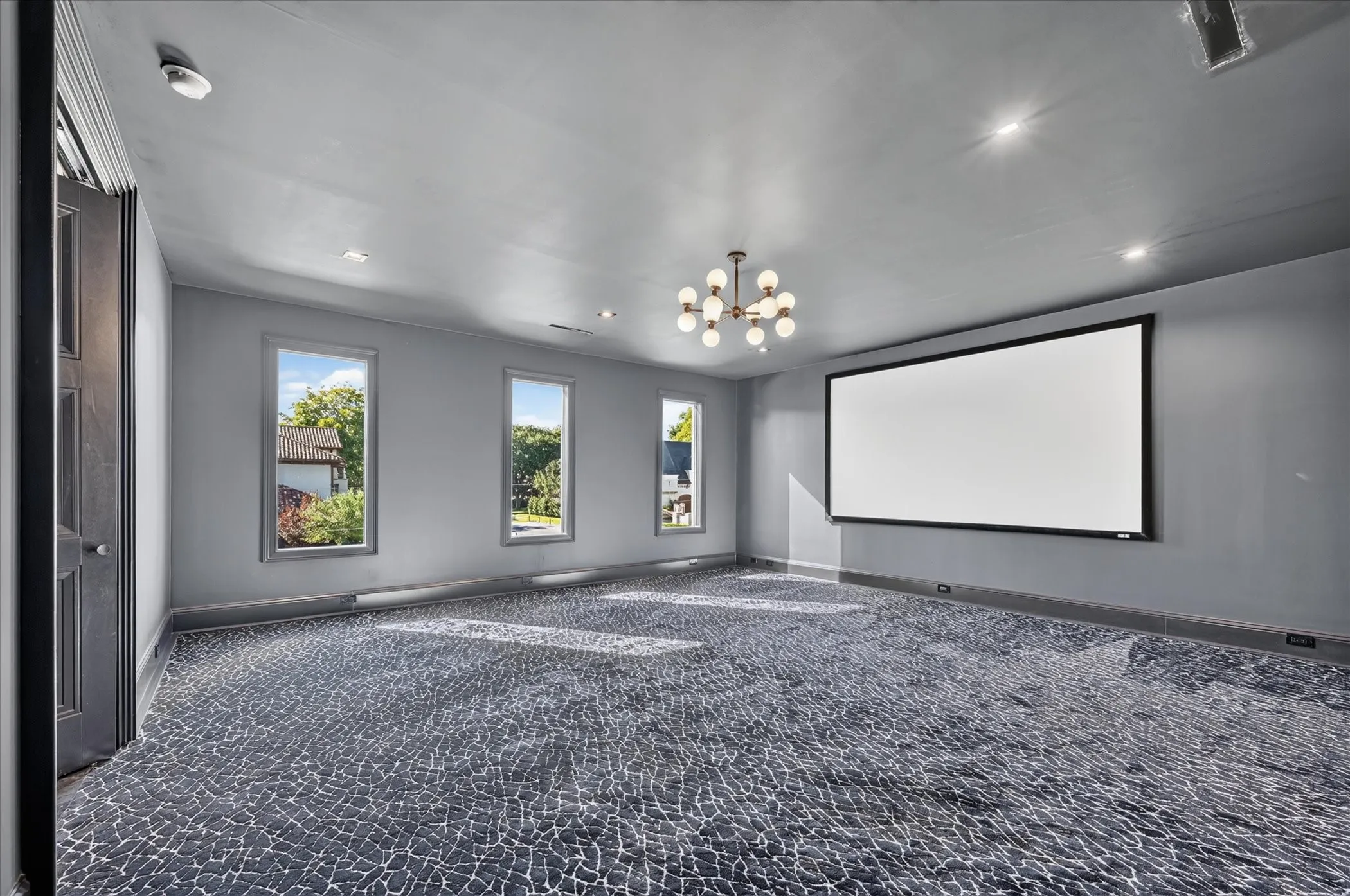 Wall-mounted projection screen, patterned wall-to-wall carpeting, contemporary chandelier, recessed lighting, and multiple windows