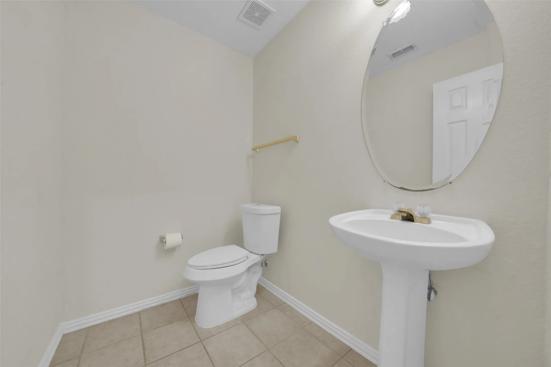 Pedestal sink with gold-tone hardware, oval wall mirror, toilet with white porcelain finish, gold-tone towel bar, and tiled flooring