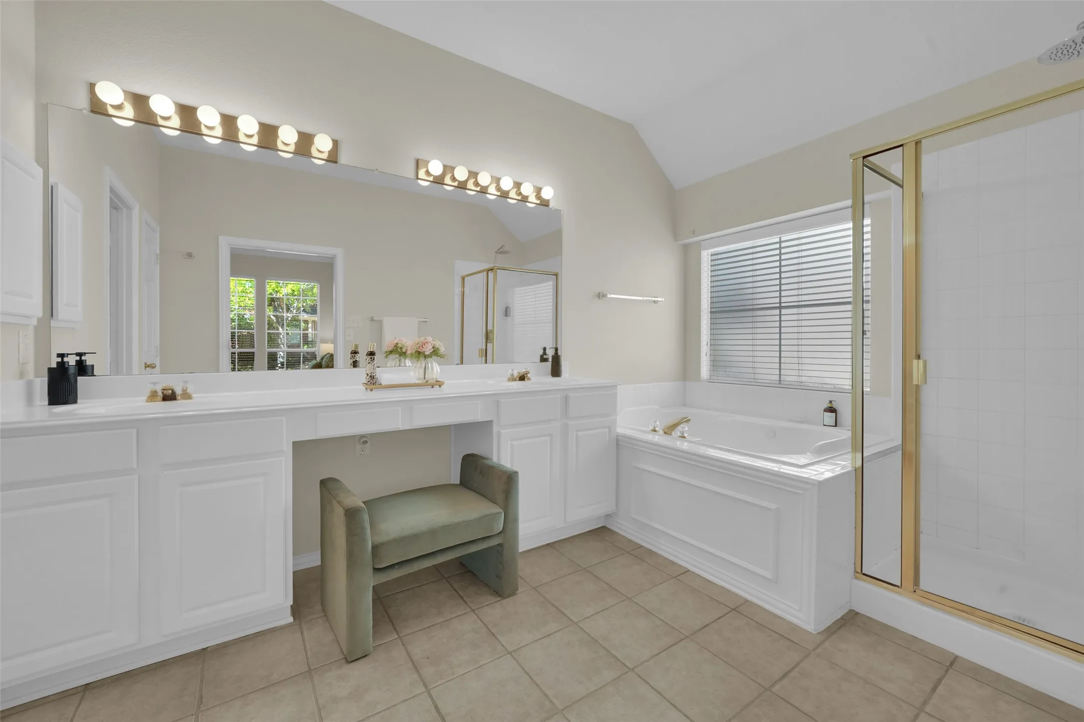 Double vanity with white cabinetry, built-in makeup counter, large vanity mirror with strip lighting, built-in soaking tub, and a glass-enclosed shower with gold-tone framing