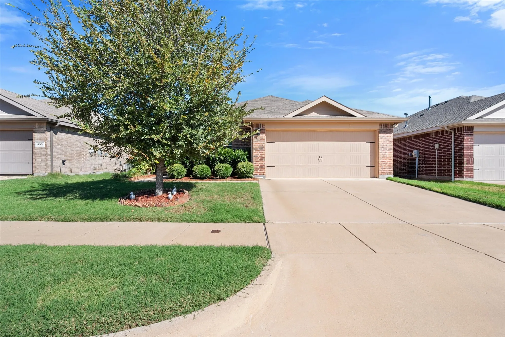Brick facade with a neutral-toned garage door, concrete driveway, and a front yard with a mature tree