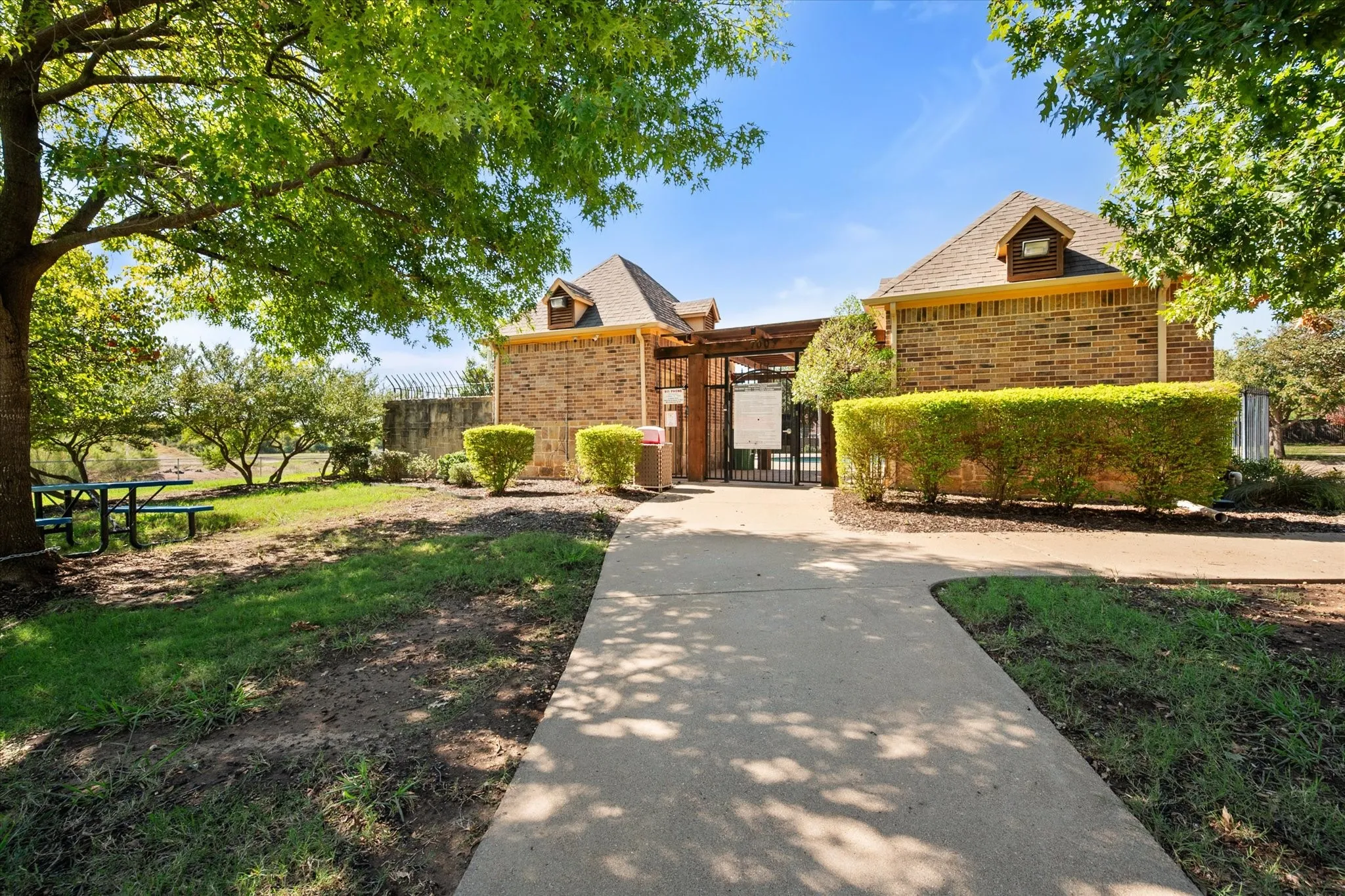 Brick-clad structure with dormer windows, surrounded by mature trees and manicured shrubbery, featuring a paved walkway and gated entry