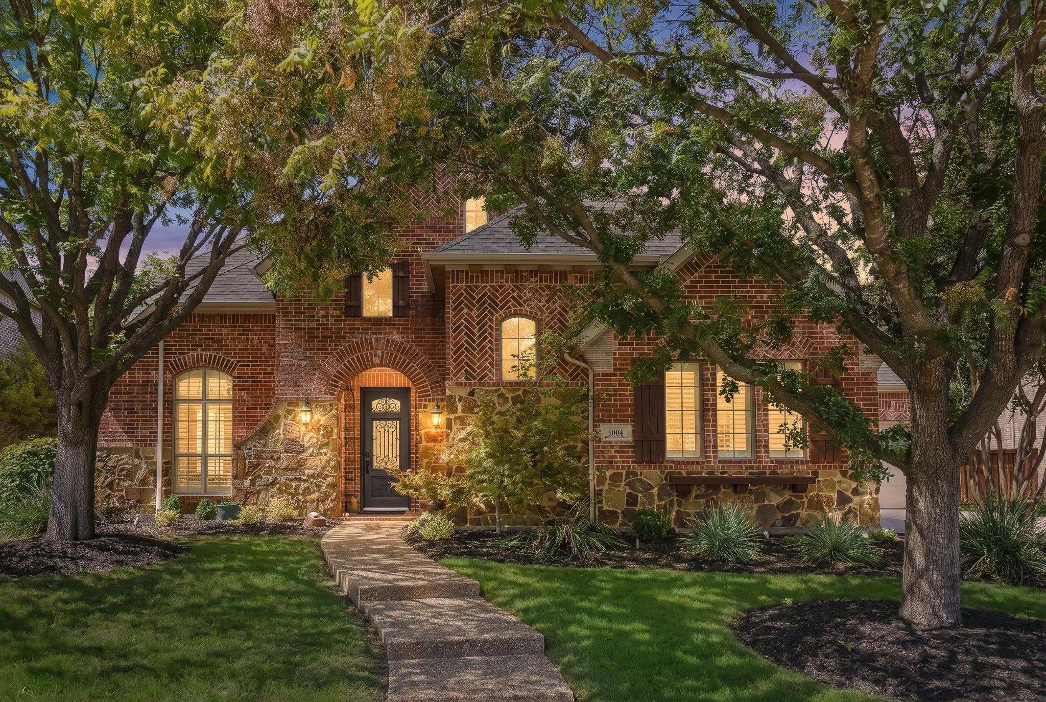 Front Exterior at Twilight | 1004 Bristlewood Drive, McKinney, Texas. Mature trees frame the illuminated facade of this five-bedroom home in Woodhaven II at Stonebridge Ranch.