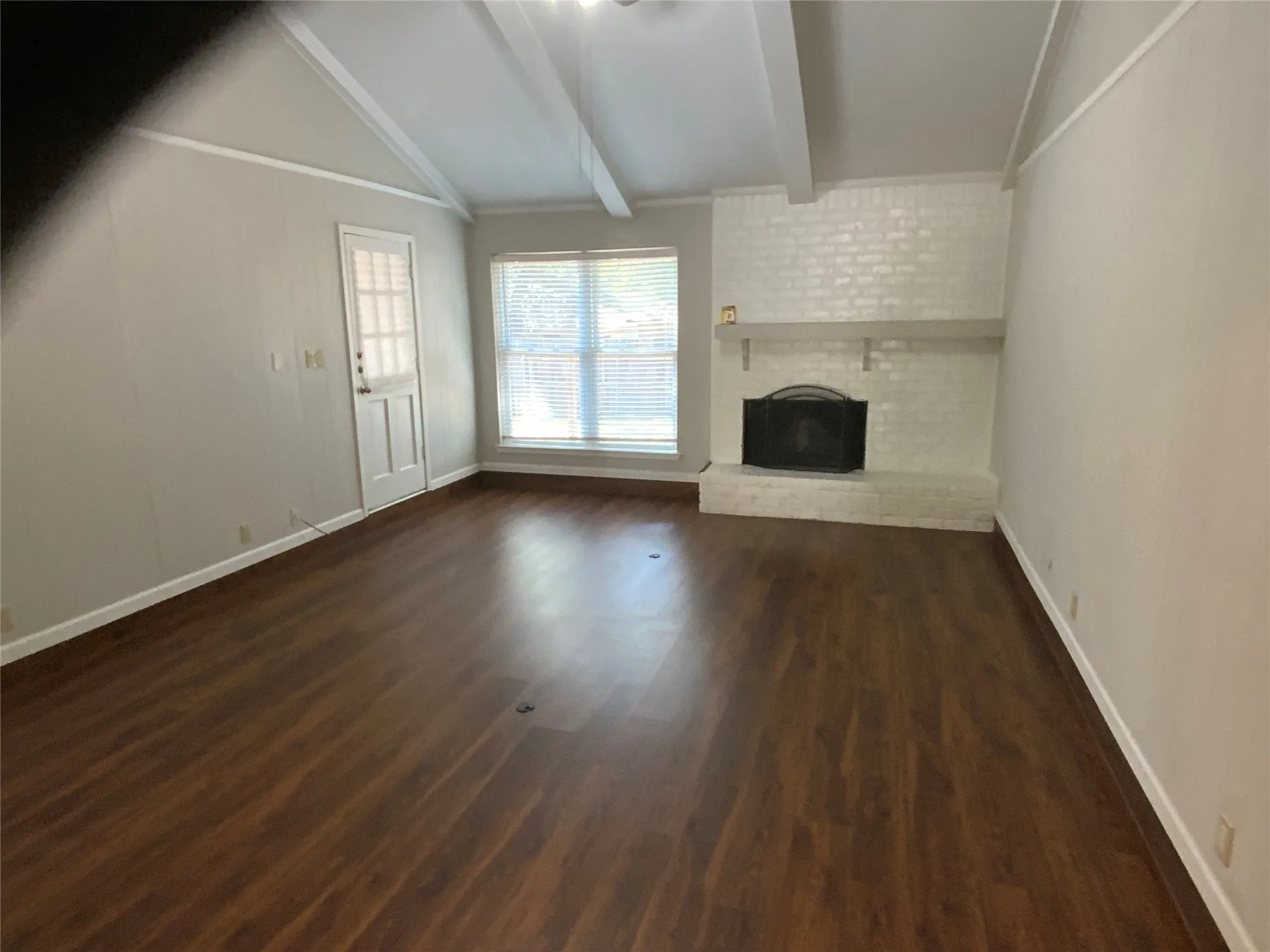 Vaulted ceiling with exposed beams, brick-faced fireplace with hearth and mantel, wood-finish flooring, and natural light from a large window