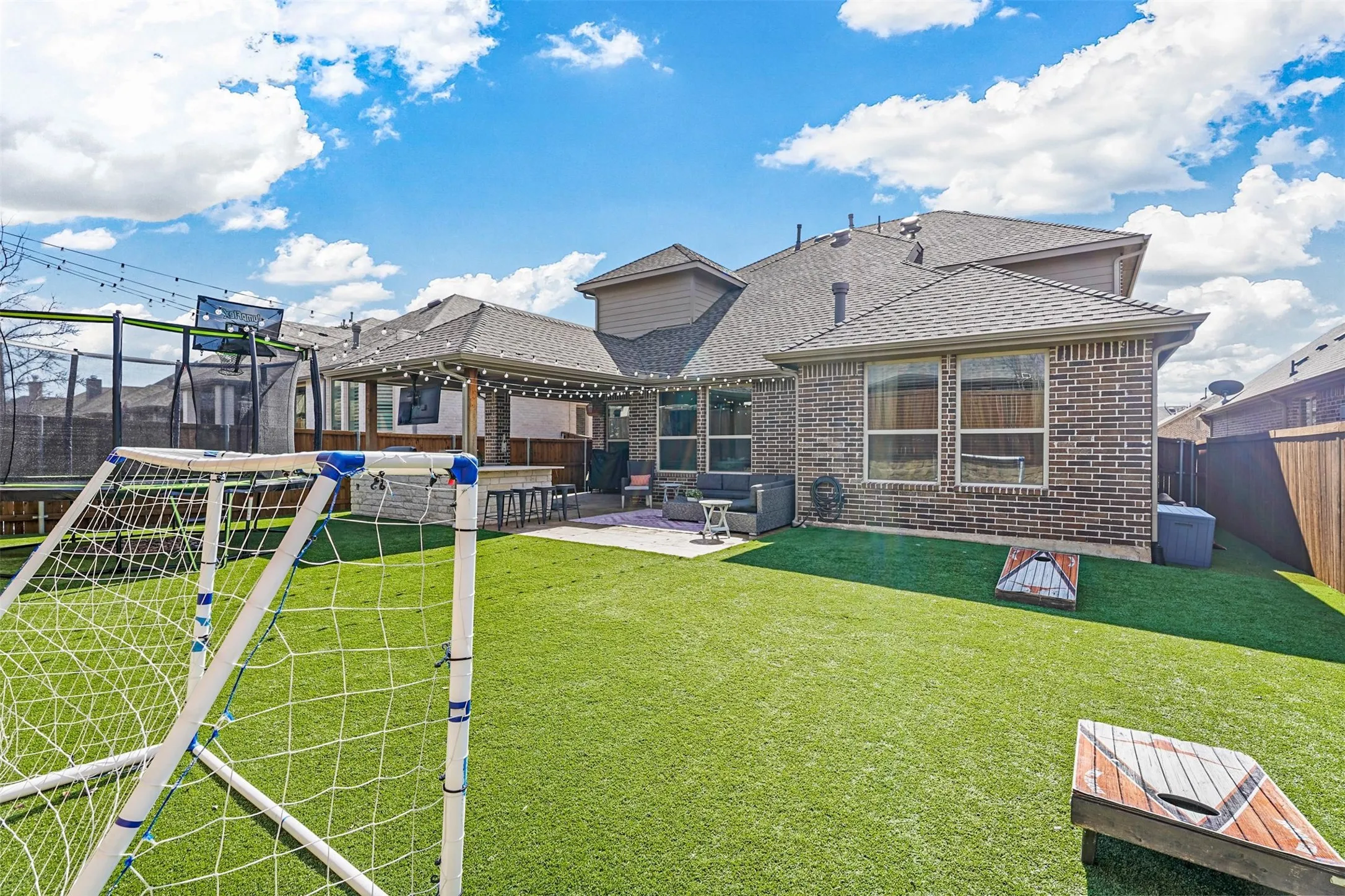 Expansive artificial turf backyard featuring a covered patio with string lights, a built-in bar, and a brick exterior