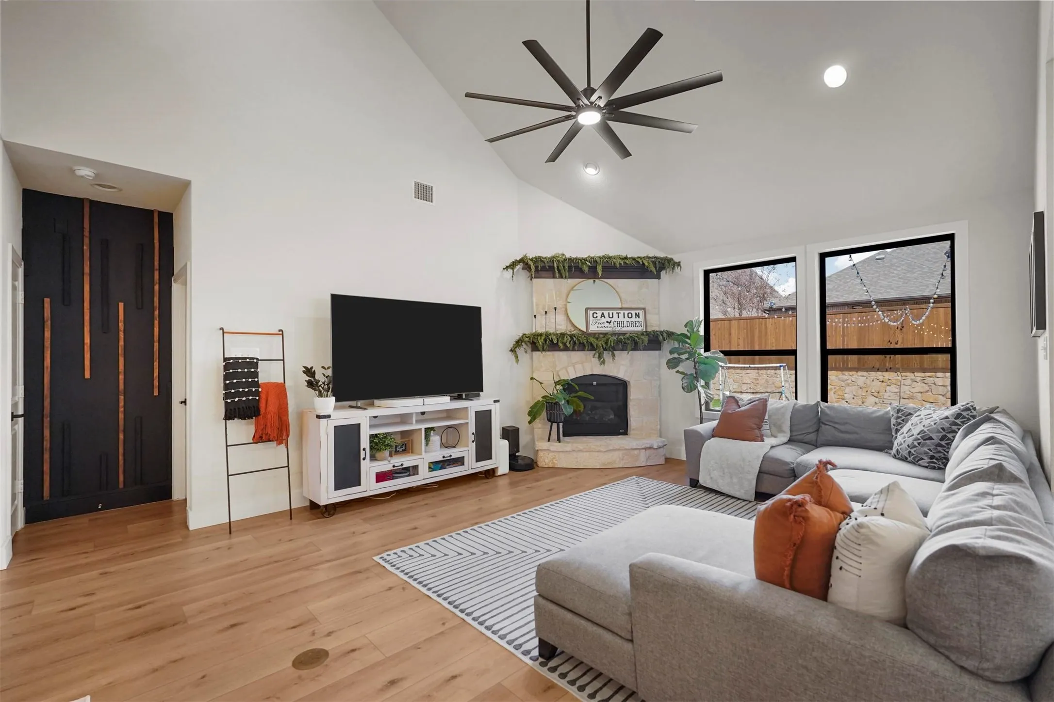 Vaulted ceiling with a ceiling fan, light wood-finish flooring, corner fireplace with a stone surround, black-framed windows, and built-in recessed lighting