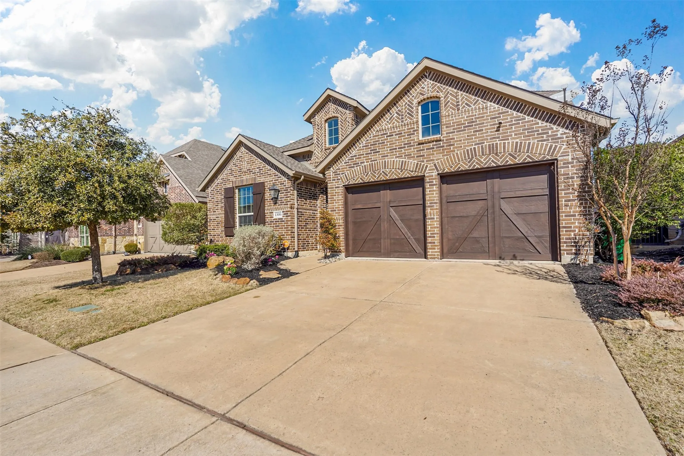 Brick exterior with arched window details, attached two-car garage with wood-finish doors, concrete driveway, and landscaped front yard with mature trees