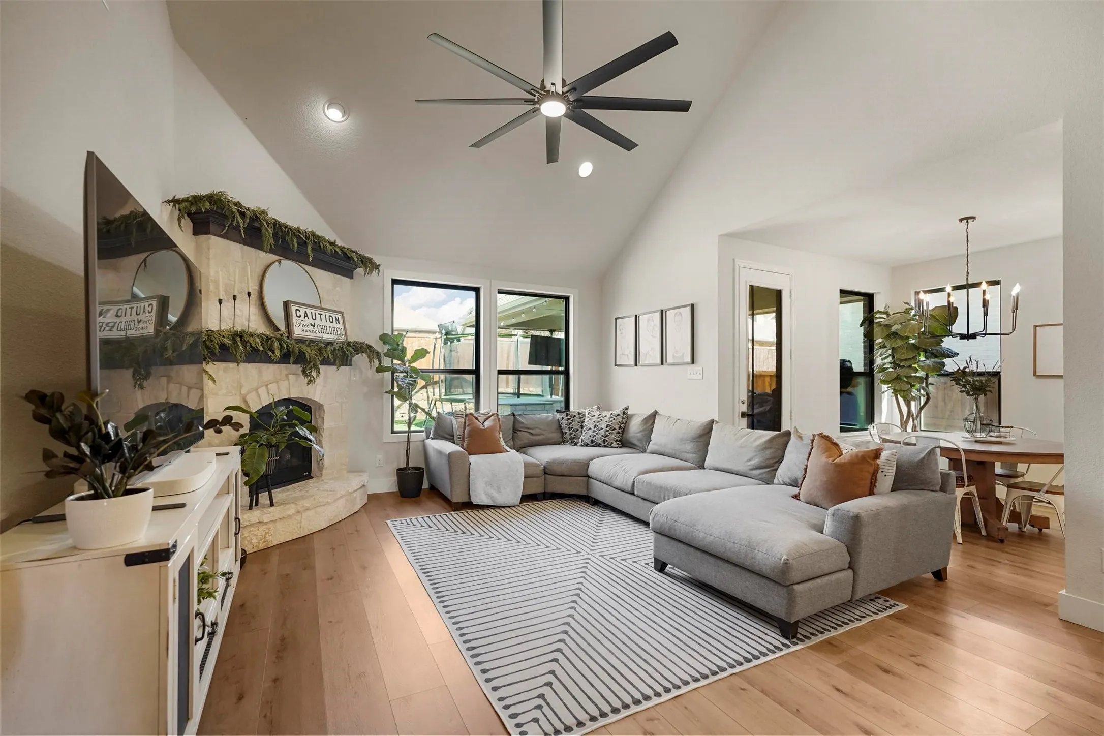 Vaulted ceiling with an integrated ceiling fan, stone-clad fireplace, large windows with black frames, and wide plank wood-finish flooring