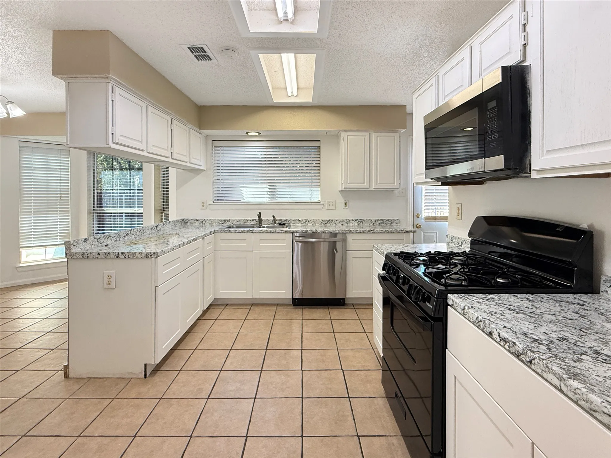 Kitchen featuring white cabinetry, light-toned tile flooring, L-shaped white stone countertops, stainless steel dishwasher, and a black range with an overhead microwave