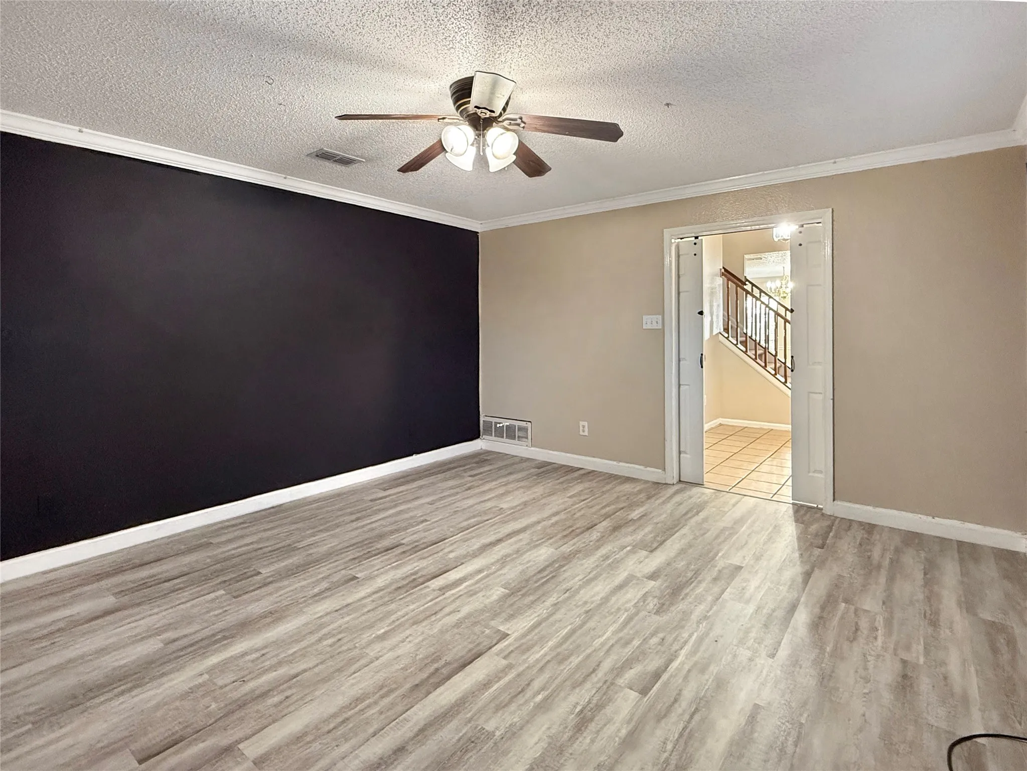 Spacious room featuring wood-finish flooring, a ceiling fan with integrated lighting, and a doorway leading to a tiled entry and staircase with metal railings