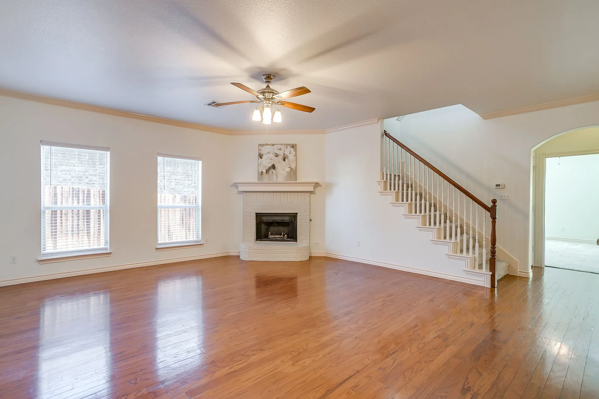 Wood-finish flooring, corner fireplace with a brick surround, white painted mantel, staircase with wood handrail, and an arched entry