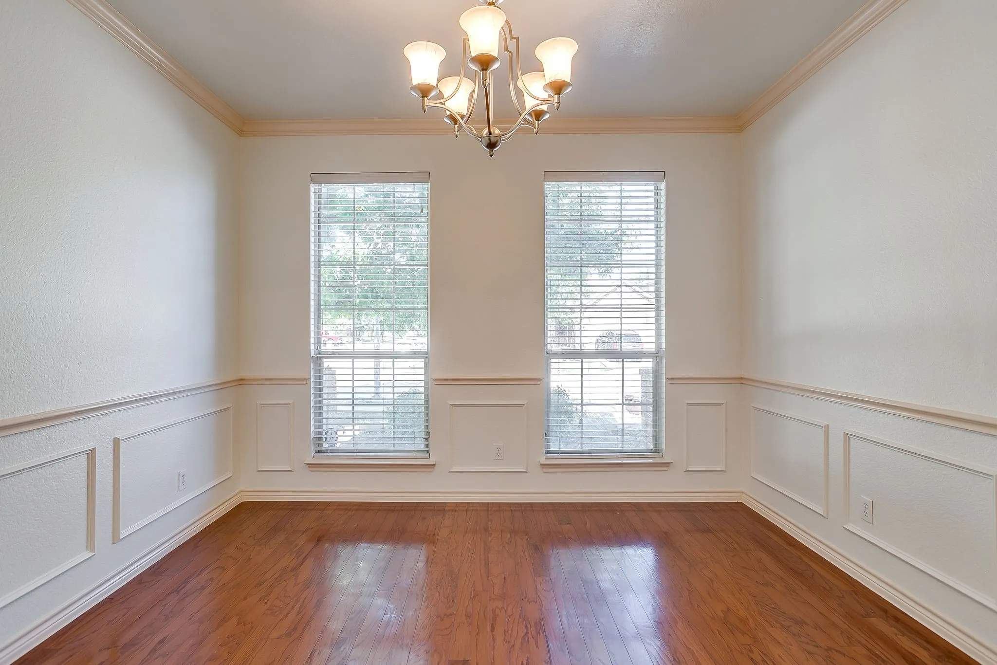 Wood-finish flooring, wainscoting, two large windows with blinds, a ceiling-mounted chandelier, and crown molding