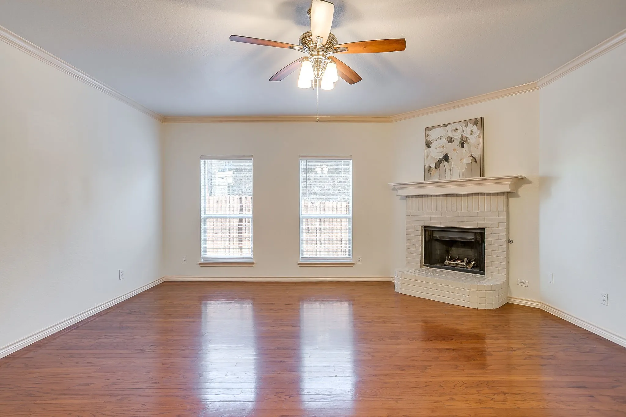 Bright interior featuring wood-finish flooring, a corner fireplace with a white brick surround and mantel, dual windows, and a ceiling fan