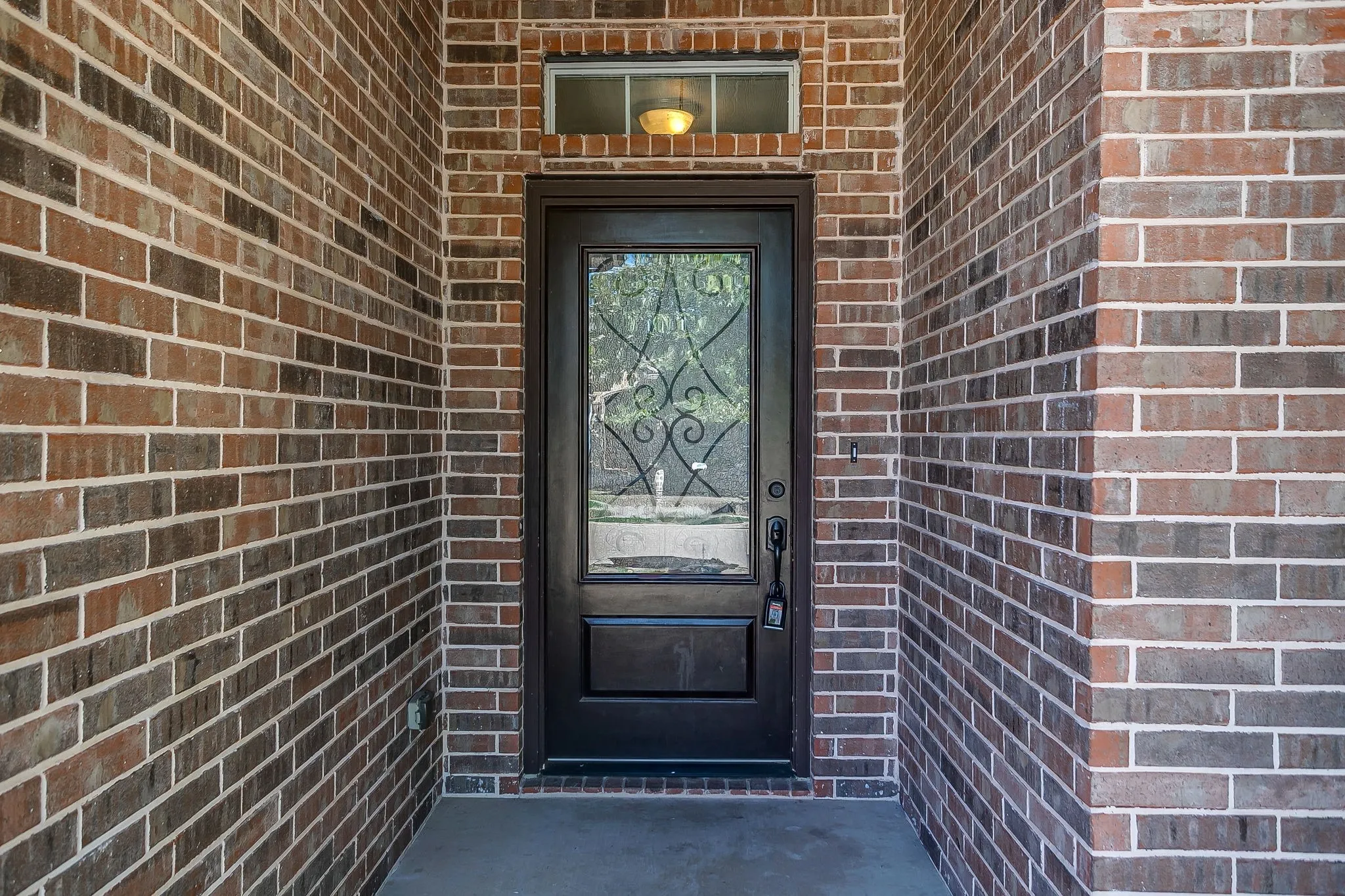 Brick exterior featuring a transom window, dark-paneled door with decorative glass inlay, and a built-in overhead light fixture