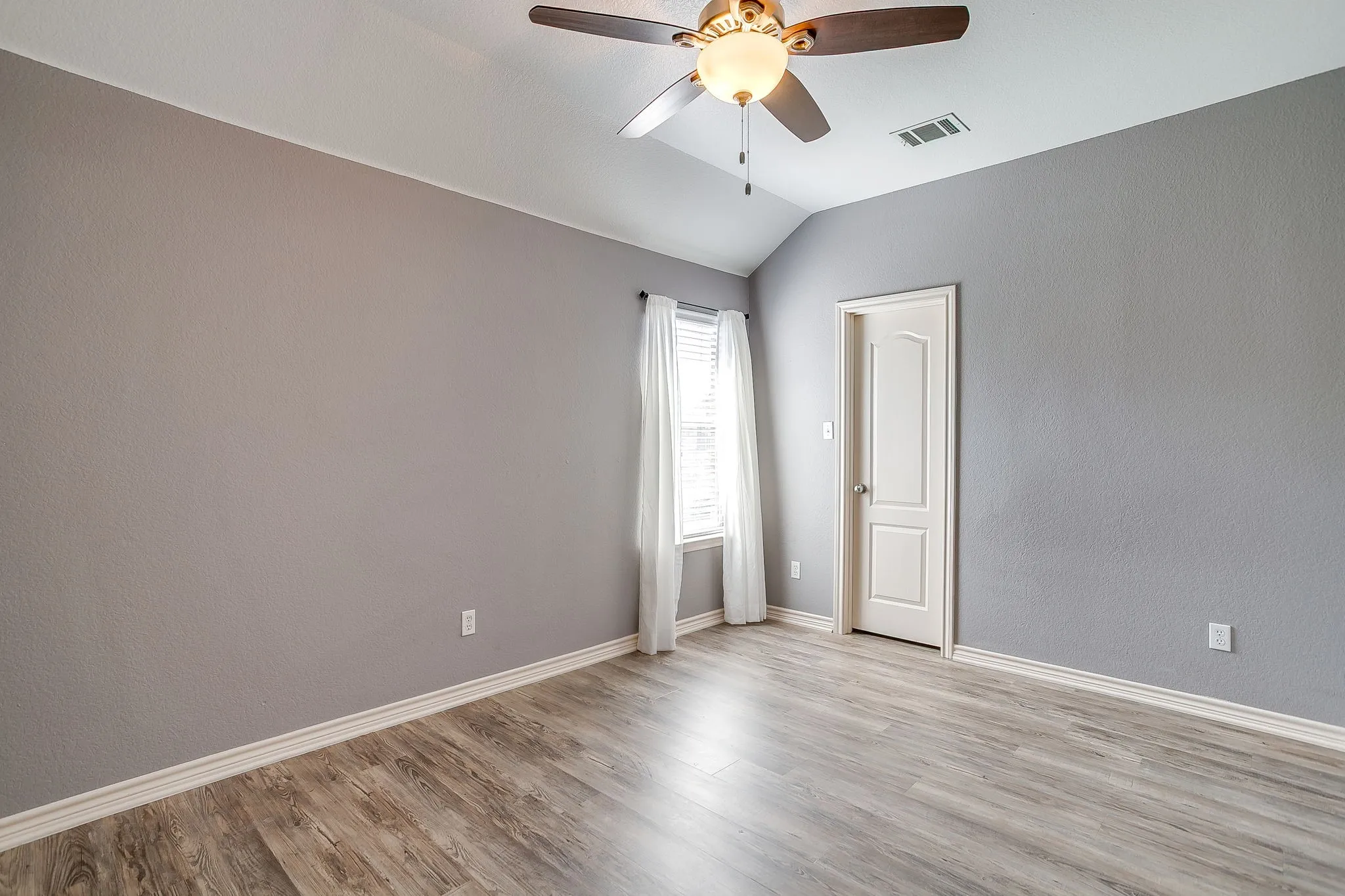 Spacious bedroom with wood-finish flooring, neutral gray walls, and abundant natural light from corner window