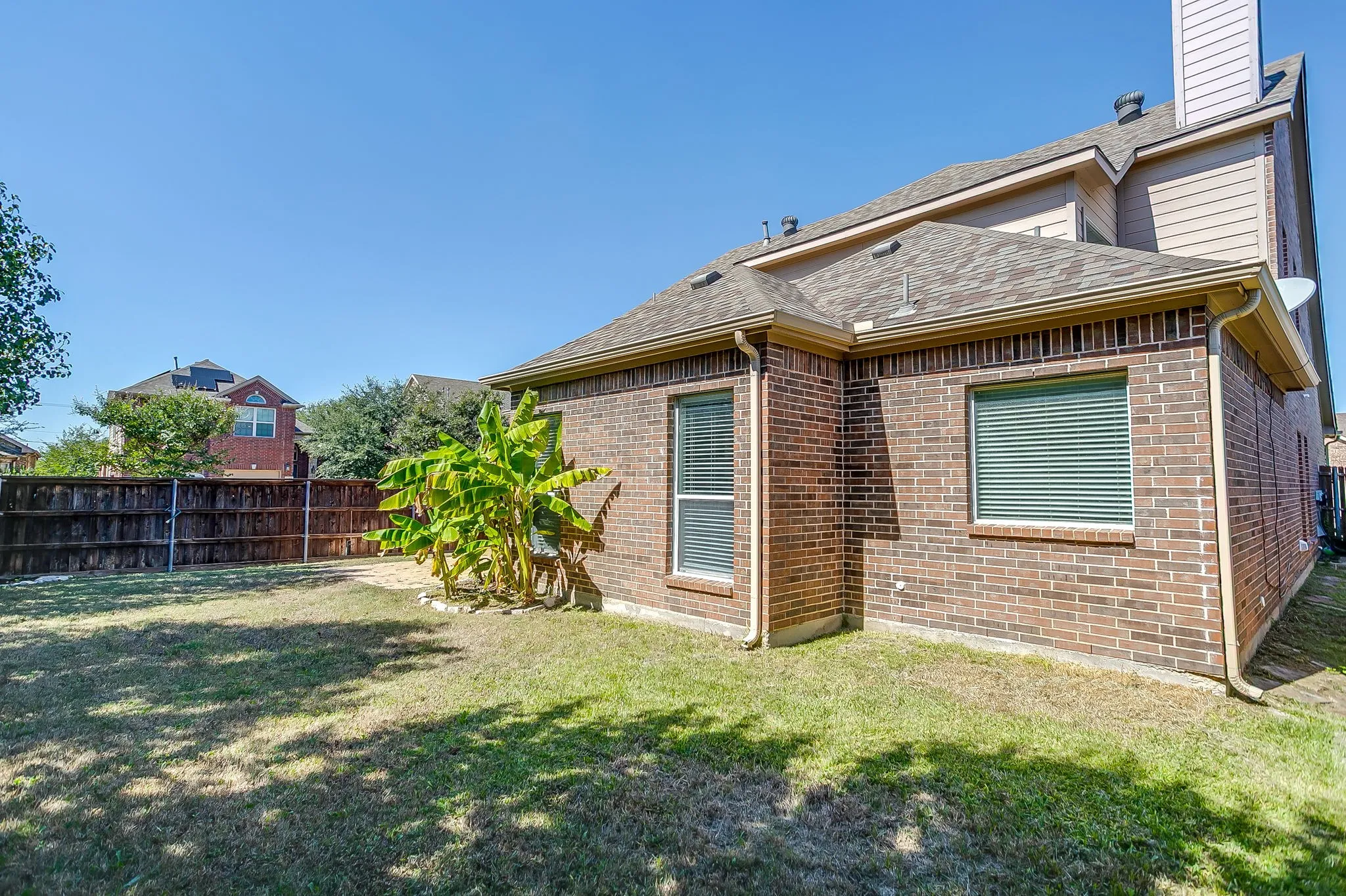 Exterior brick facade with a multi-level roofline, featuring an expansive lawn, a privacy fence, and multiple windows