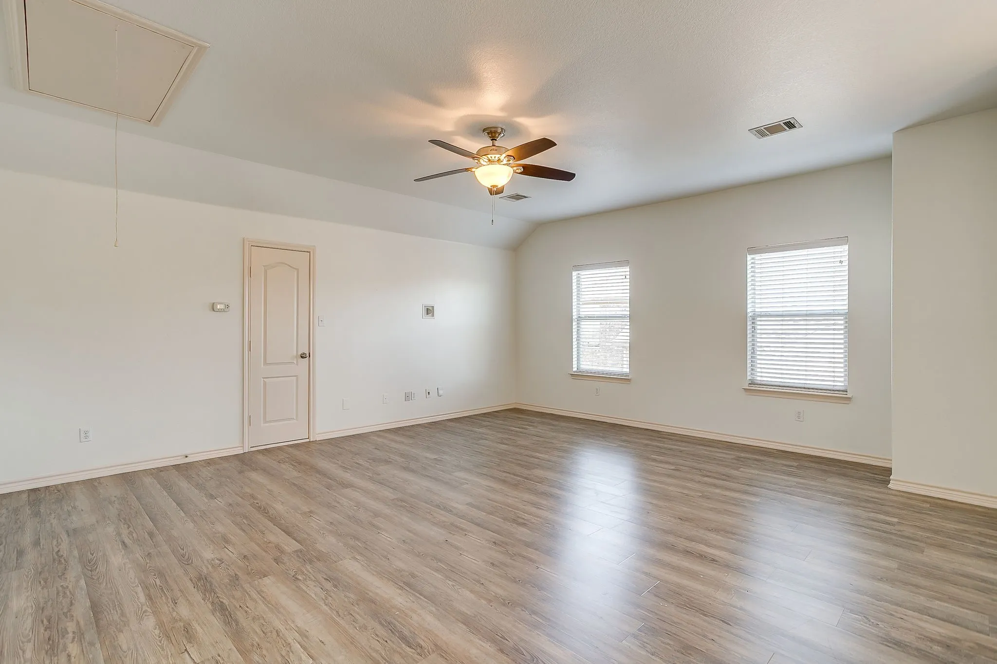 Spacious room featuring wood-finish flooring throughout, abundant natural light from dual windows with plantation shutters, ceiling fan with integrated lighting, and a neutral color palette with soft gray-toned walls