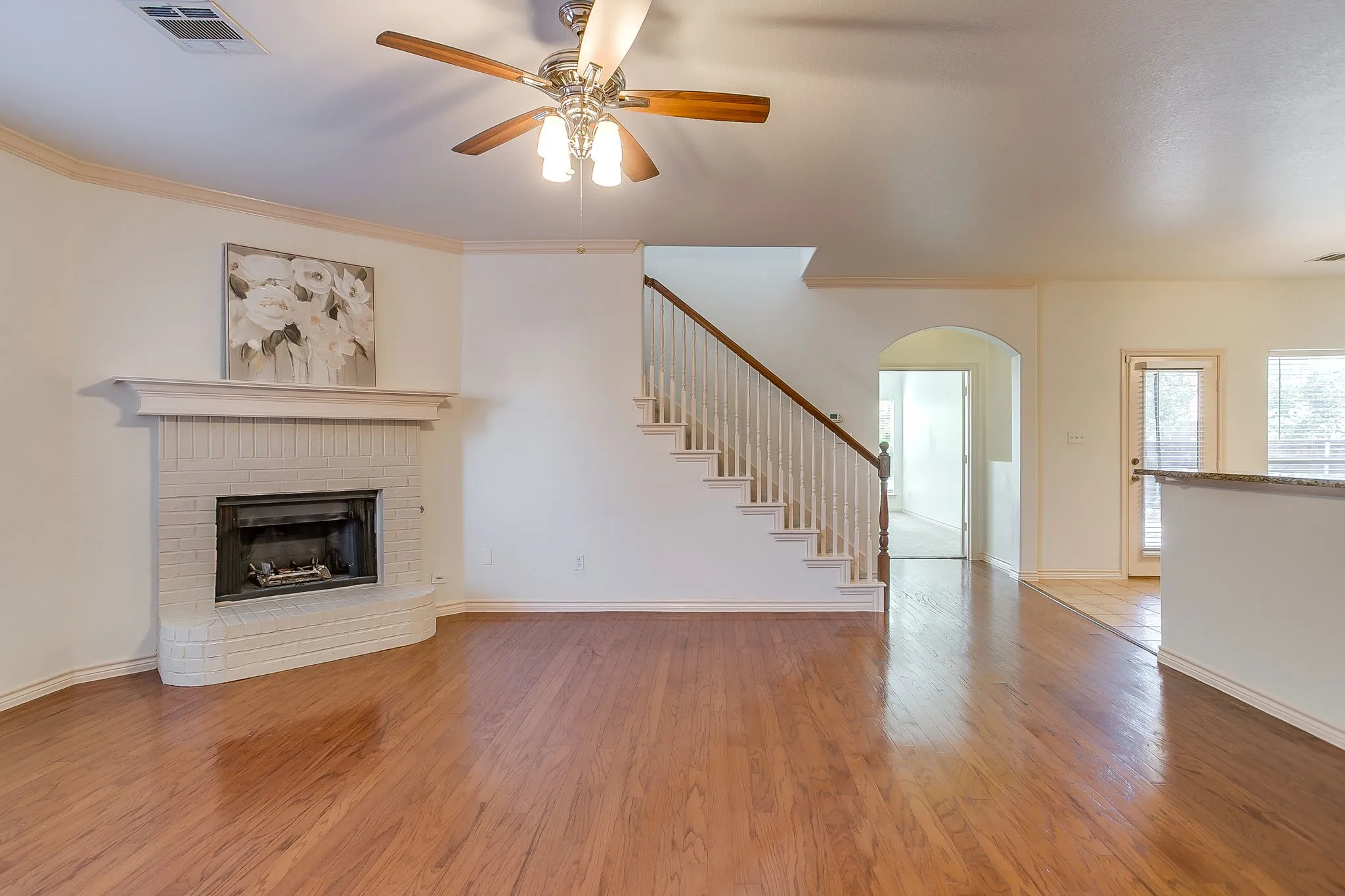 Hardwood flooring throughout, a brick fireplace with a mantle, a ceiling fan with integrated lighting, a staircase with wood handrails, and an arched doorway