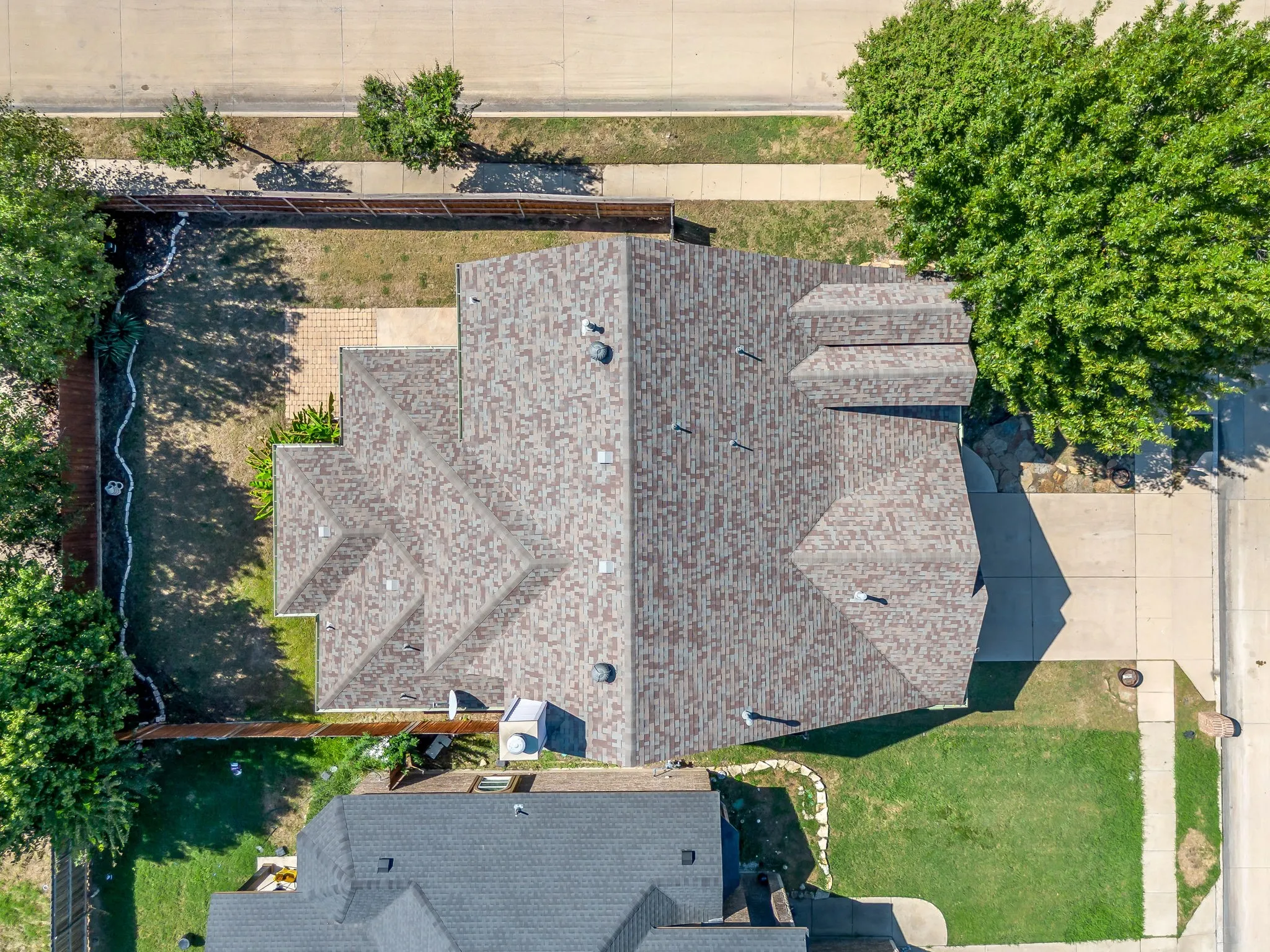 Aerial view showcasing a residential property with a newly installed asphalt shingle roof, expansive manicured lawn, mature landscaping with established trees along the perimeter, and defined hardscaped areas