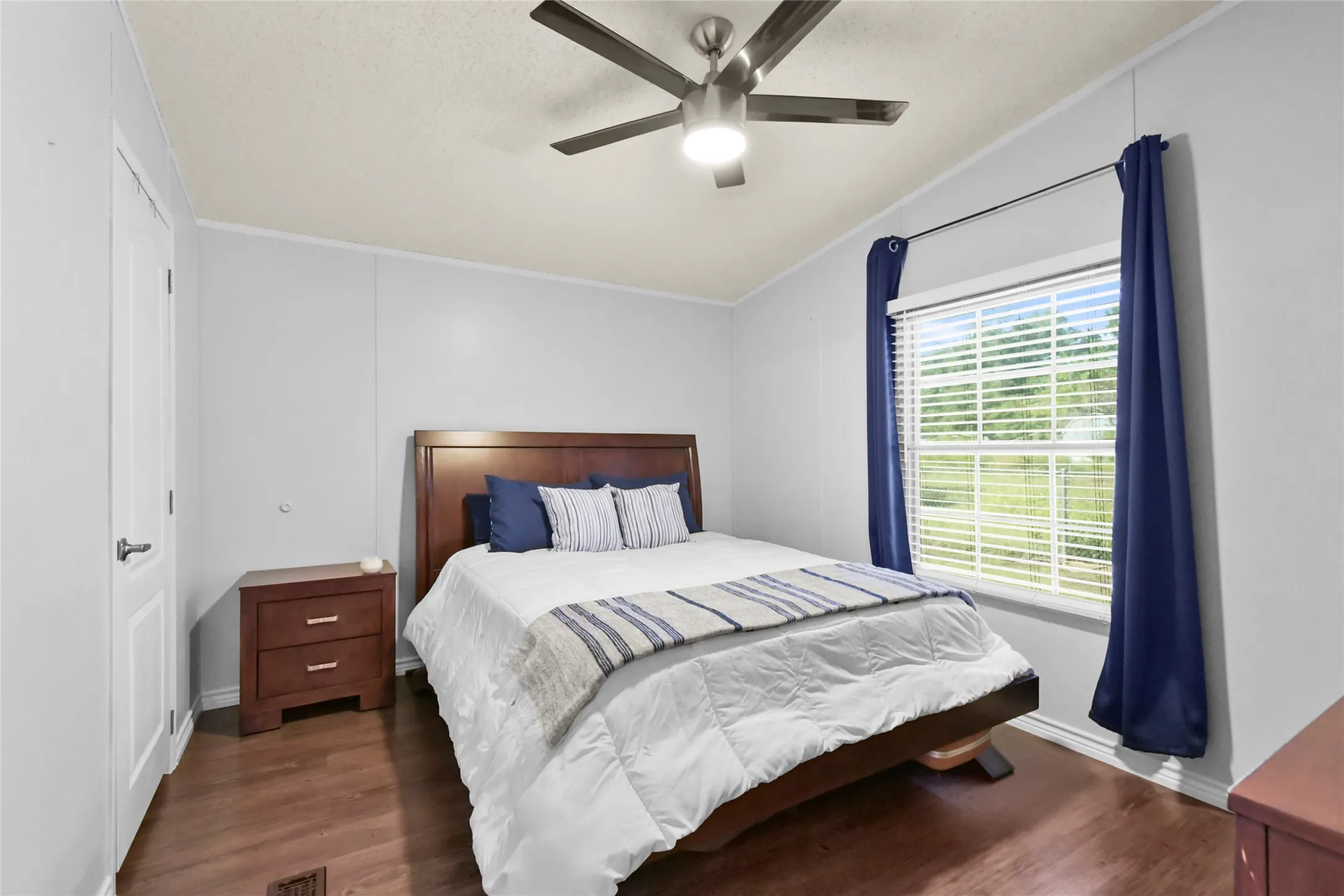 Bright secondary bedroom featuring wood-finish flooring, a ceiling fan with integrated lighting, large window with blue curtain panels providing natural light, wood nightstand, and white painted walls with crown molding detail
