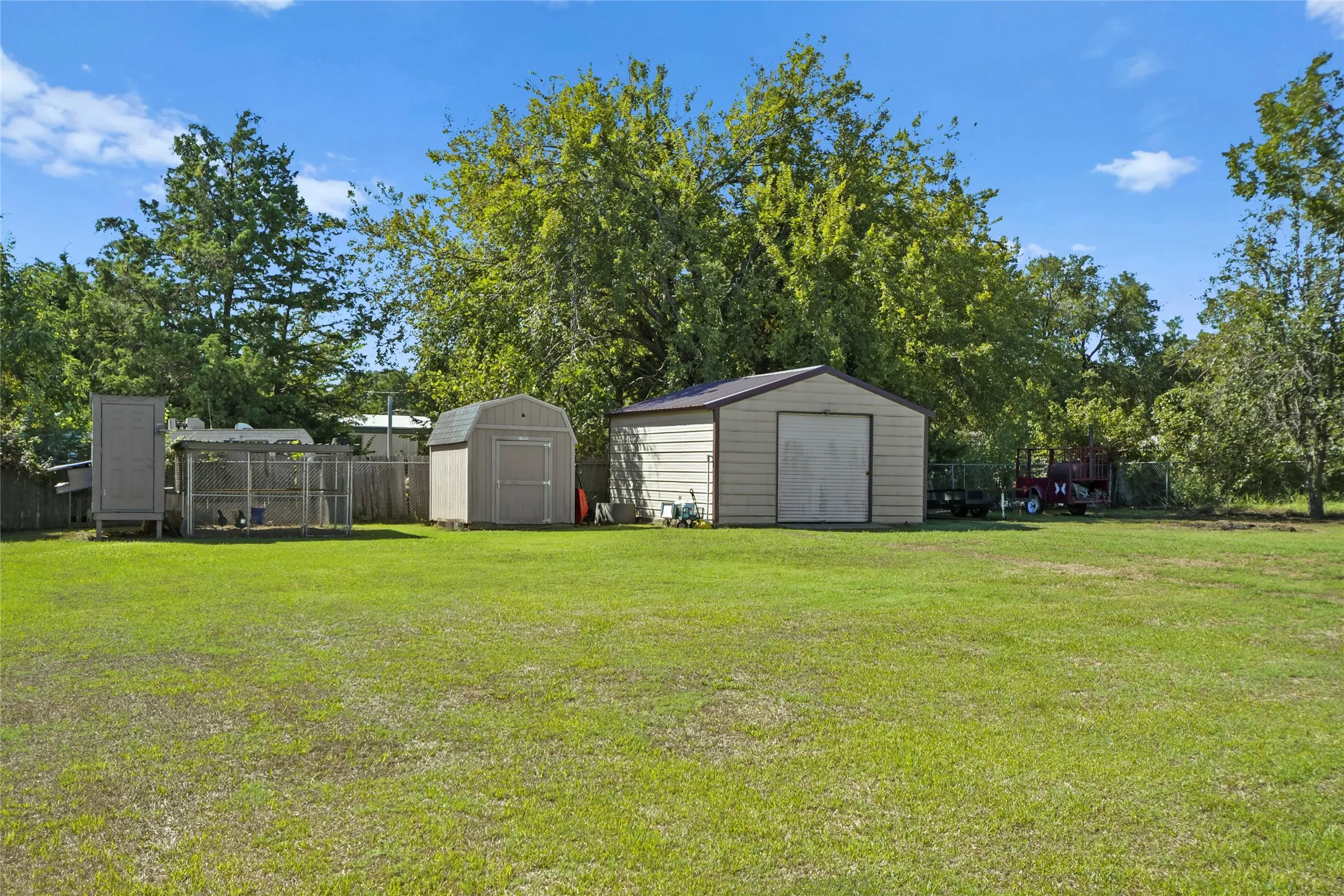 Expansive green space featuring two storage sheds, a fenced enclosure, and mature trees