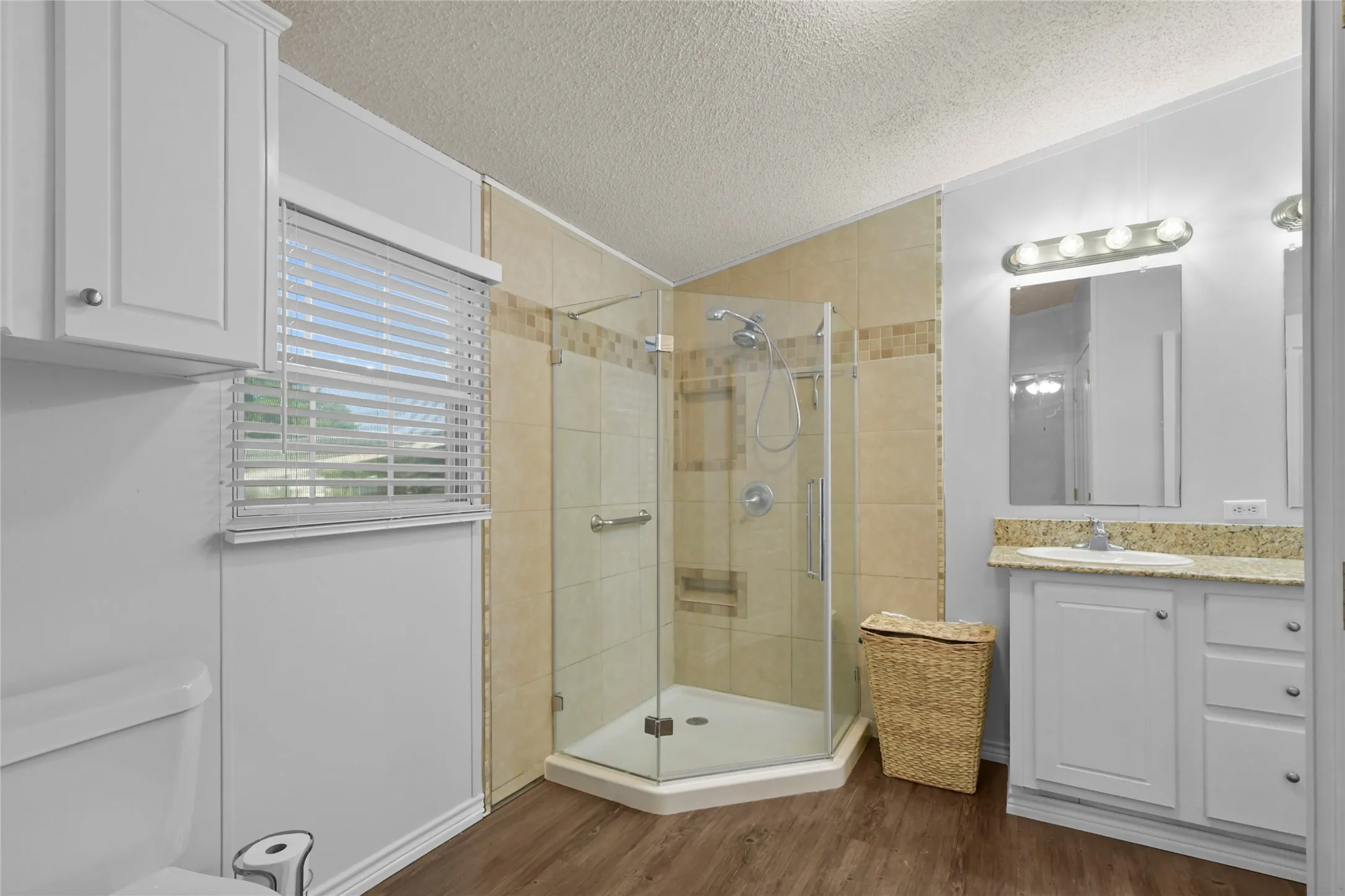 Shower enclosure with glass panels and tile surround, single vanity with a granite-style countertop, wood-finish flooring, and white cabinetry