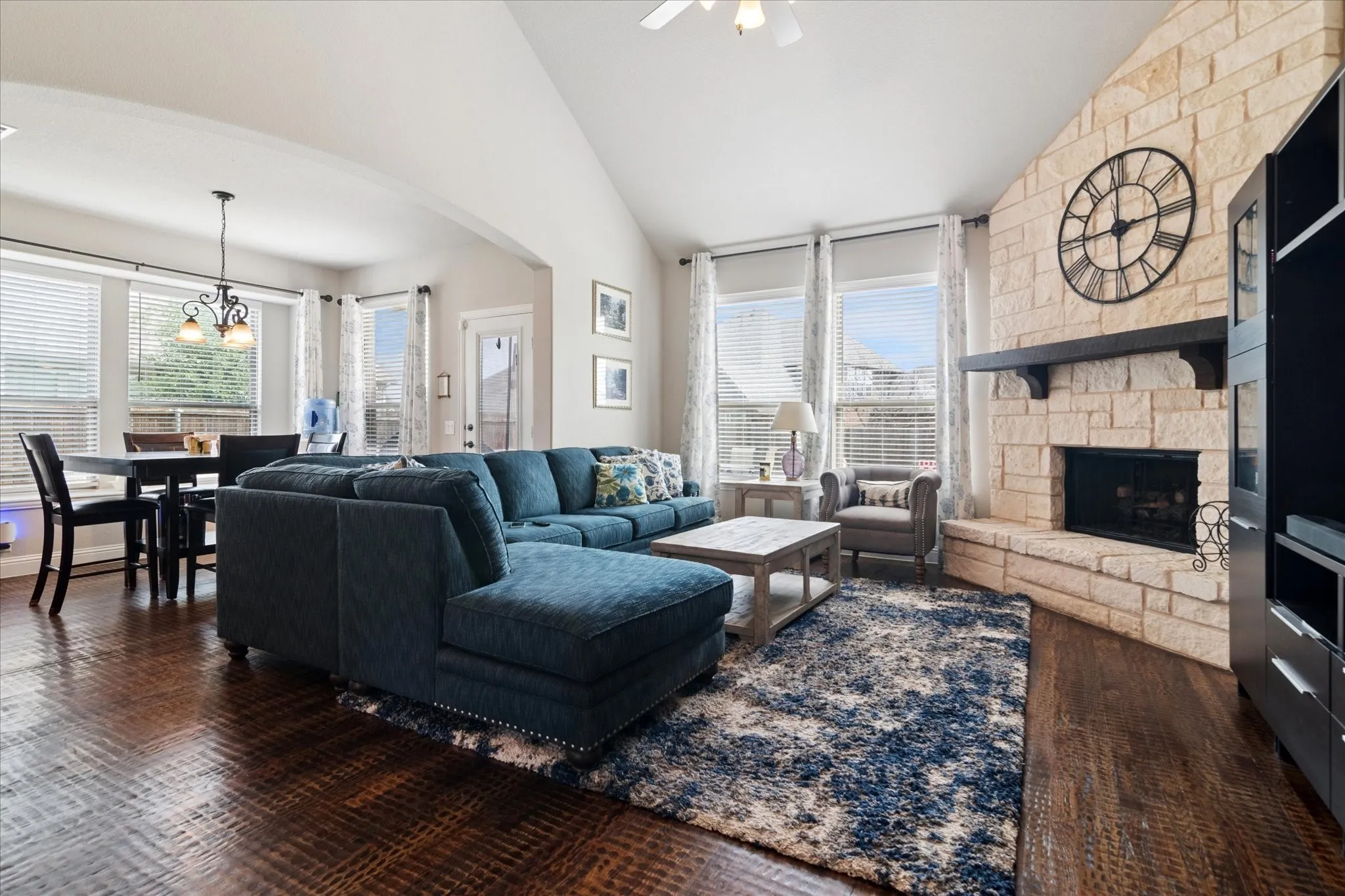 Stone-clad fireplace with a dark wood mantel, high vaulted ceiling, dark wood-finish flooring, and multiple windows with blinds
