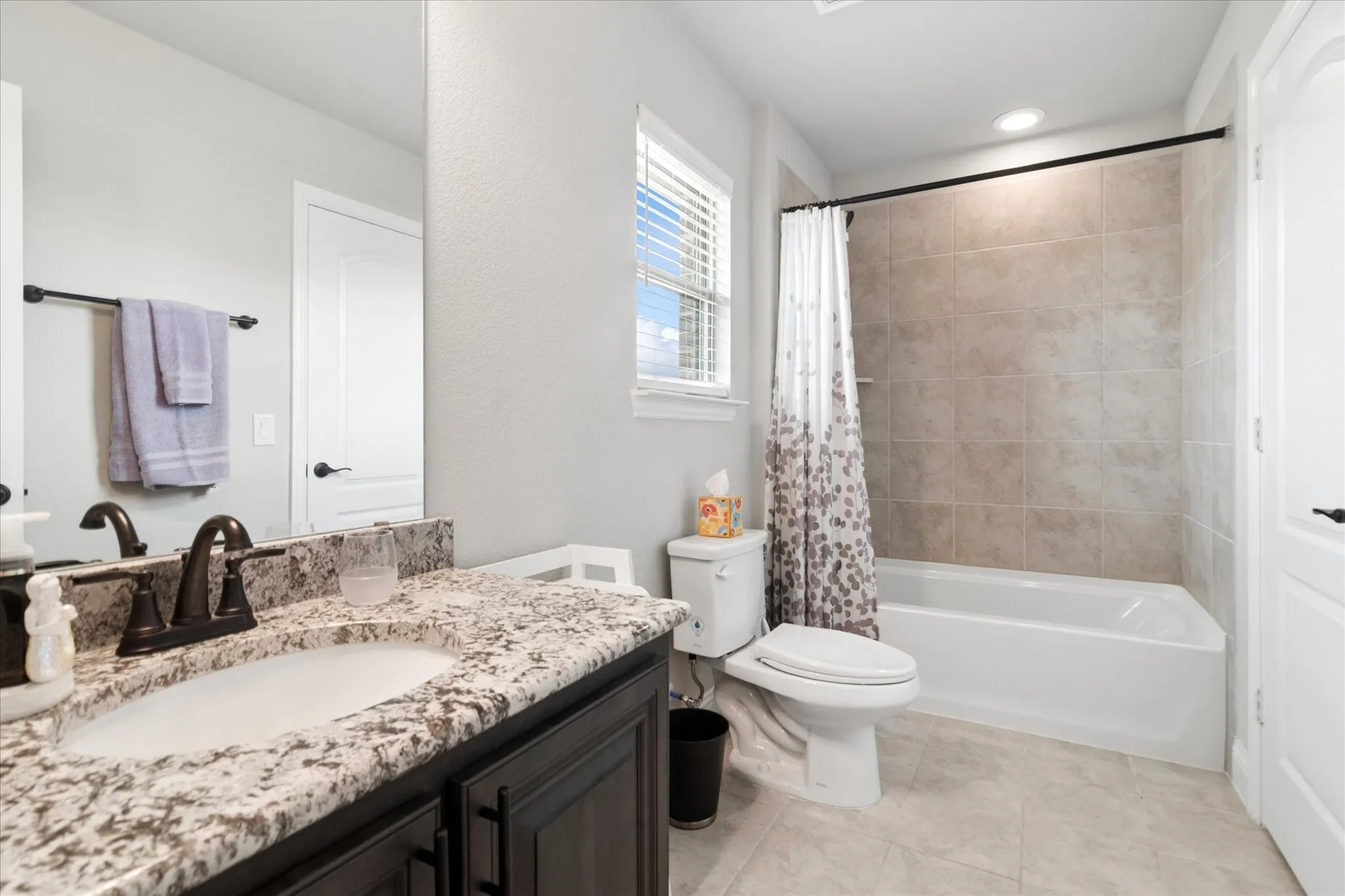 Vanity with granite countertop and undermount sink, dark wood-finish cabinetry, bathtub with tiled shower surround, recessed lighting, and tile flooring