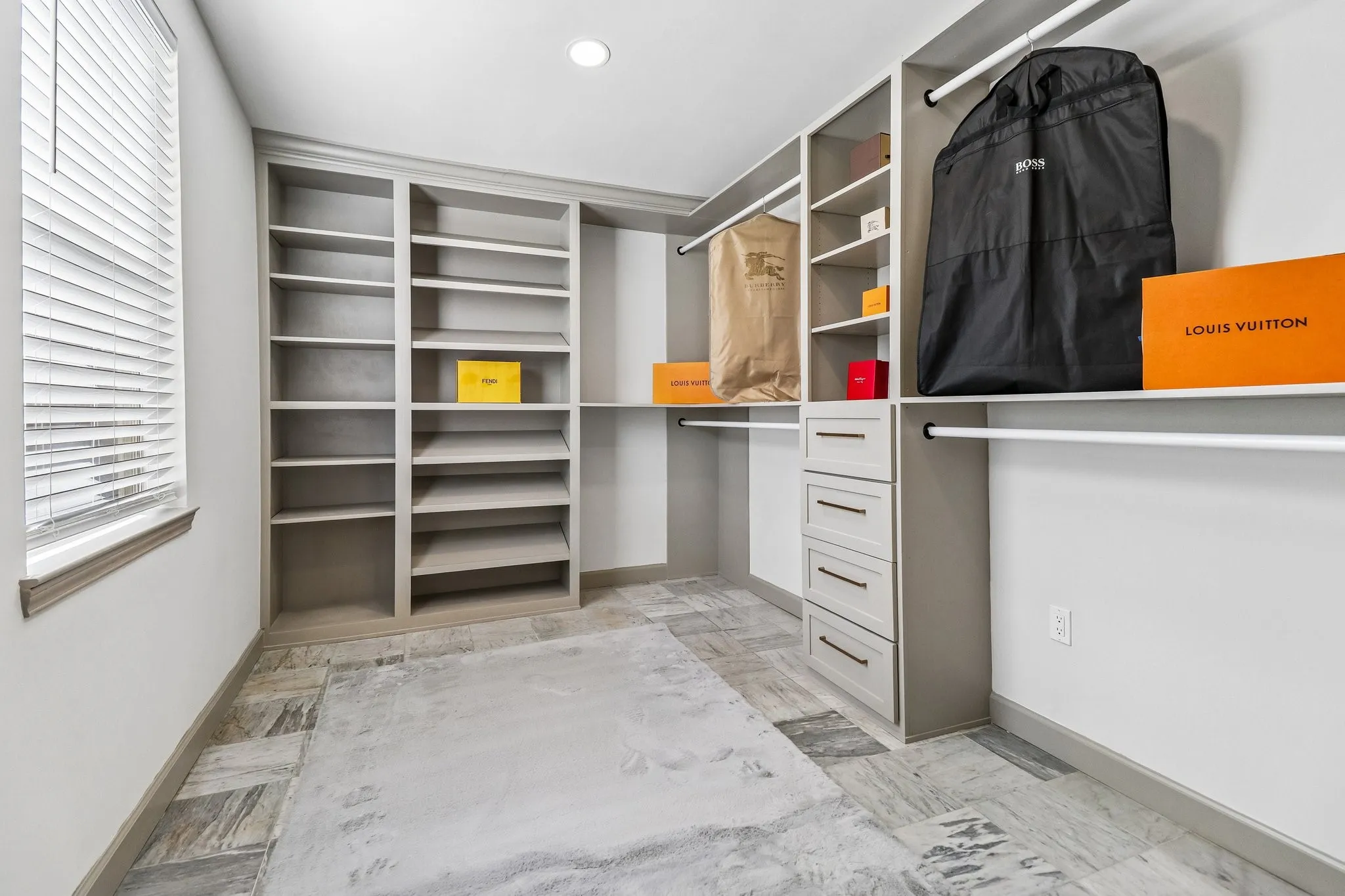 Custom built-in shelving, integrated drawers, white hanging rods, window with blinds, and varied stone-look floor tiles