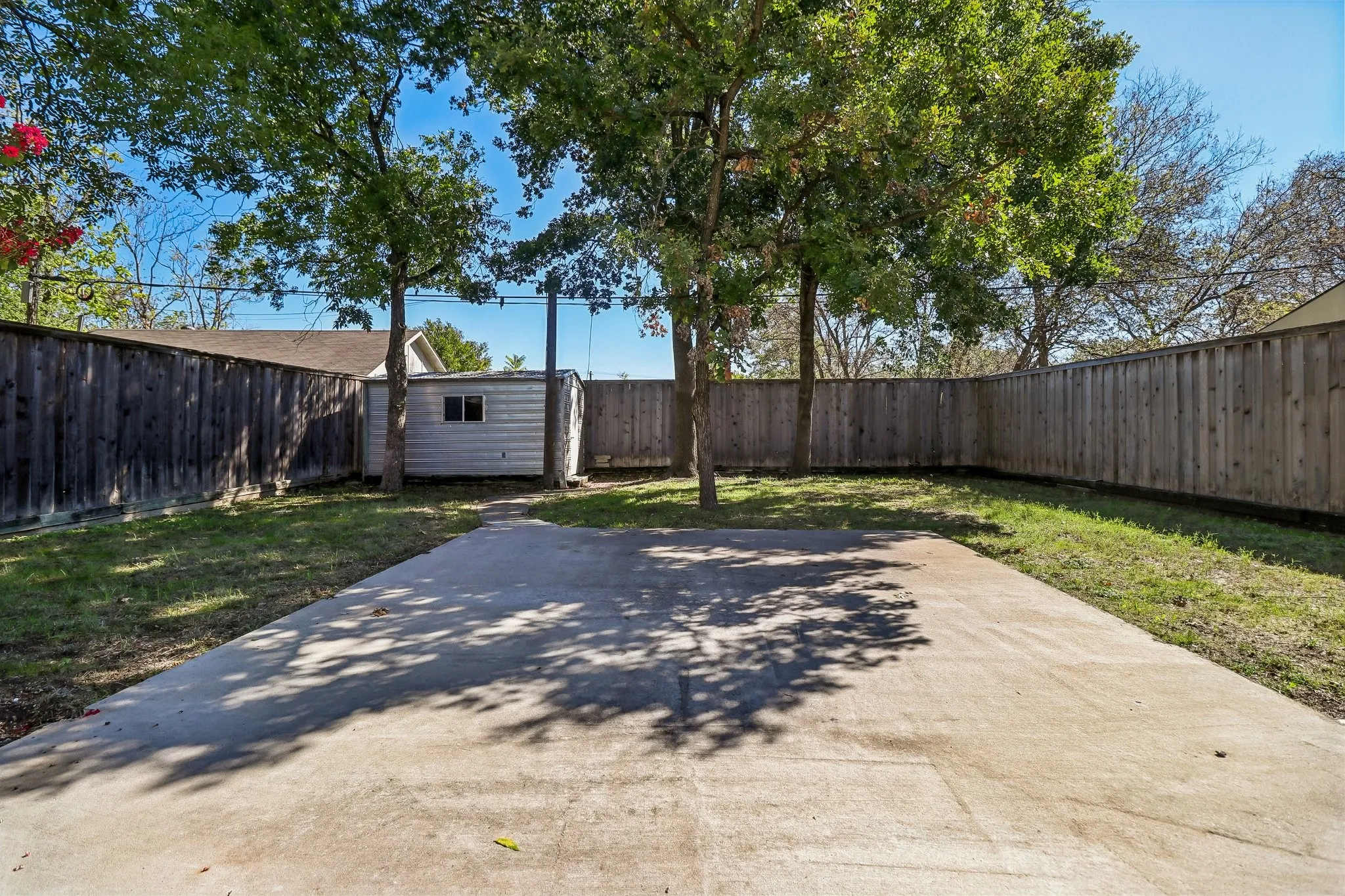 Expansive concrete pad, lush lawn, mature trees, wood privacy fencing, and a detached storage shed