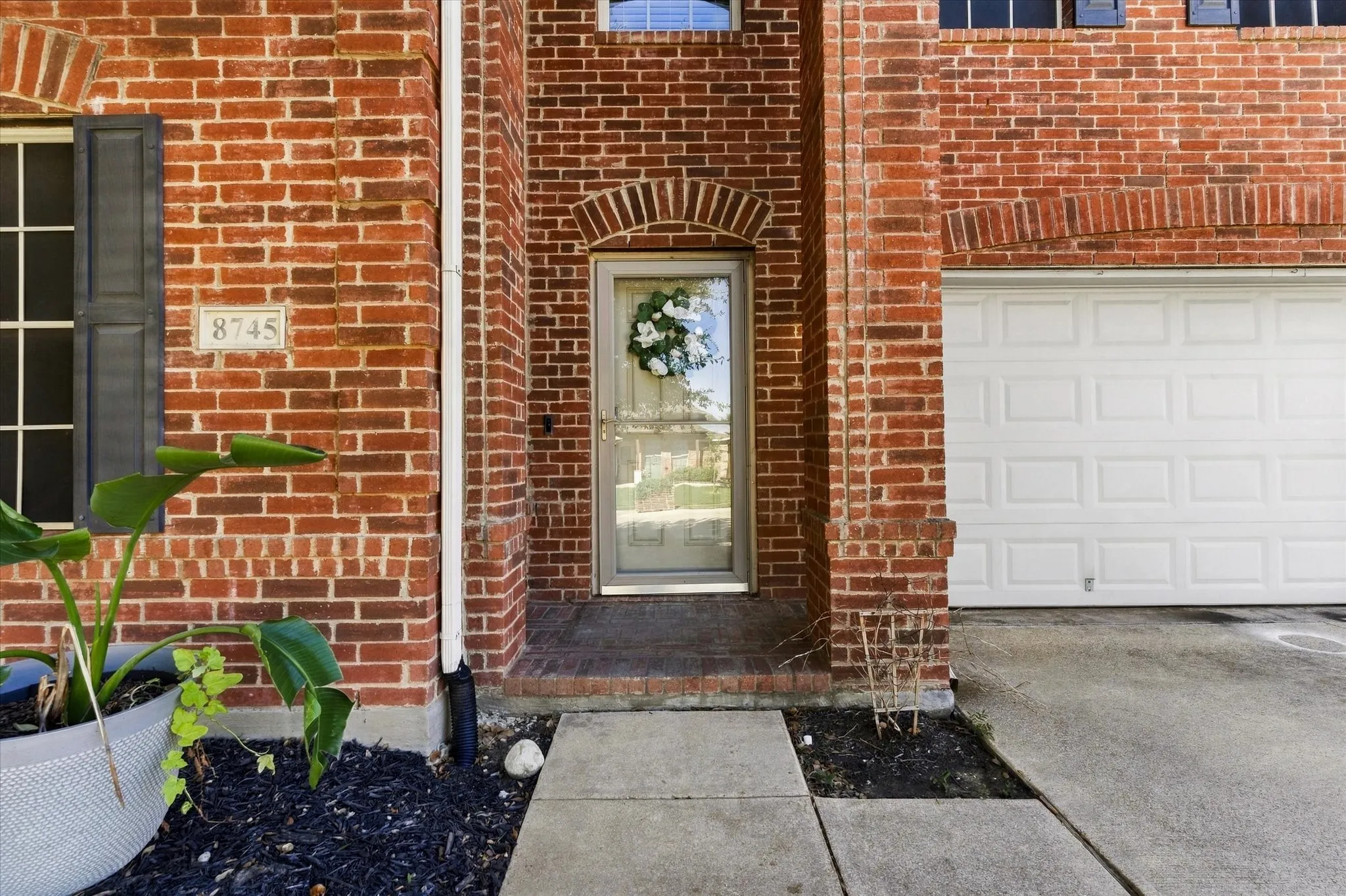 Red brick exterior, arched doorway, panel-style garage door, concrete walkway, and landscaped planting beds