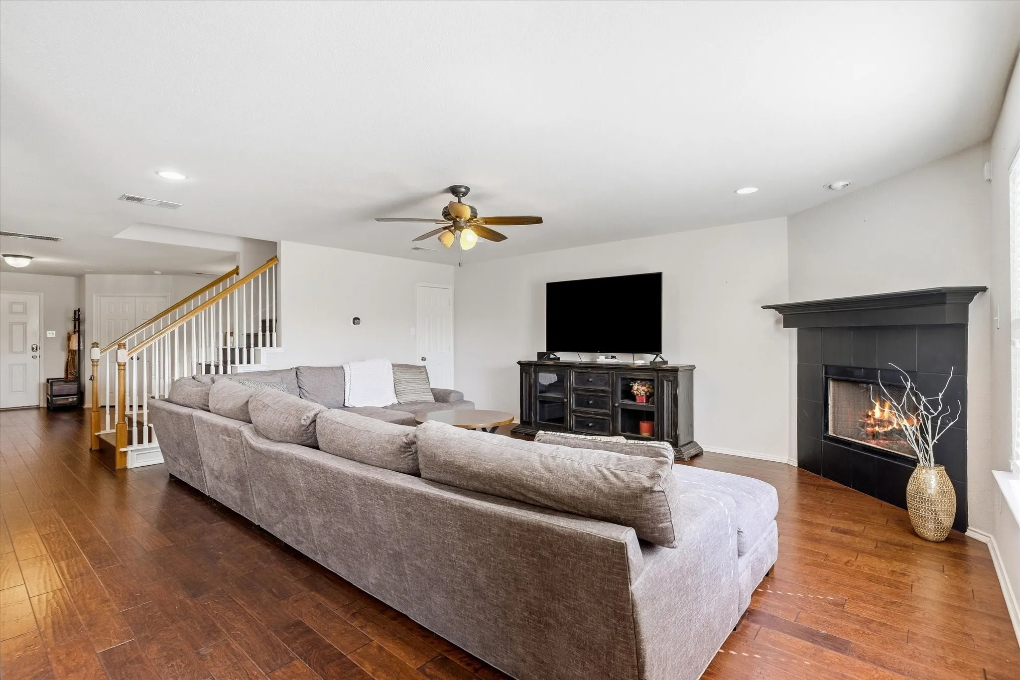 Wood-finish flooring throughout, corner fireplace with dark mantel, white painted walls, ceiling fans, and a staircase with wood handrails and white balusters