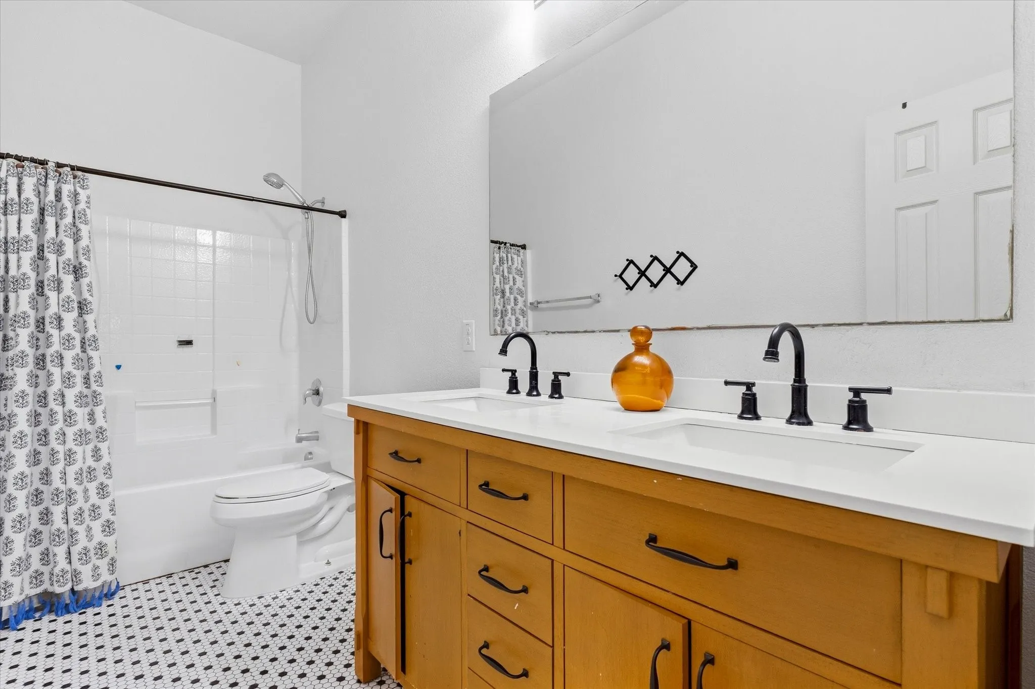 Wood-finish double vanity with dual sinks, matte black hardware, large wall-mounted mirror, and black and white floor tiling